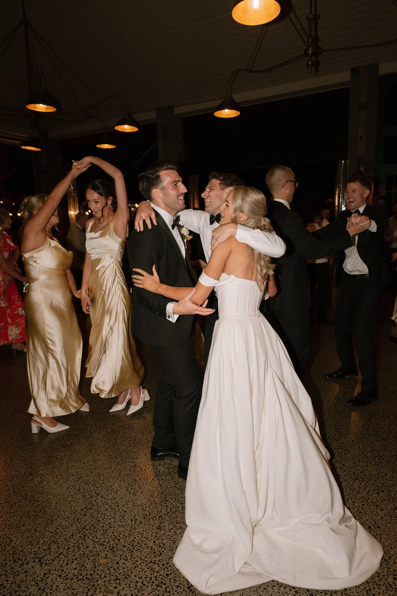 Wedding reception with couples dancing, including bride in white gown and groom in tuxedo, under warm lighting.