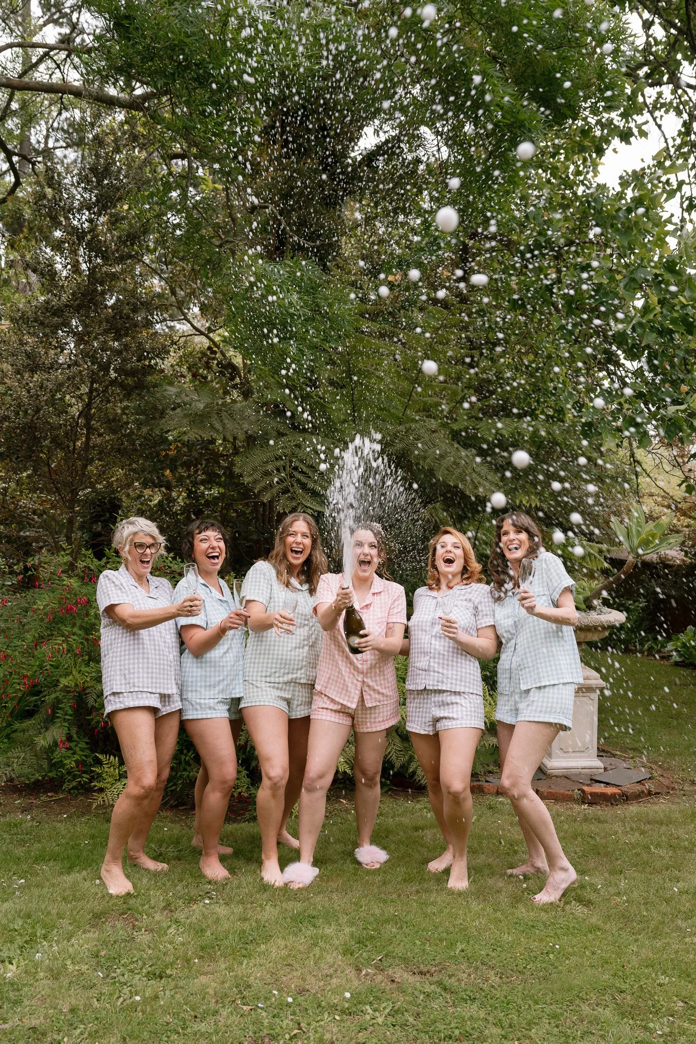 Group of six women in pajamas celebrating outdoors as they spray champagne, smiling and enjoying in a garden.
