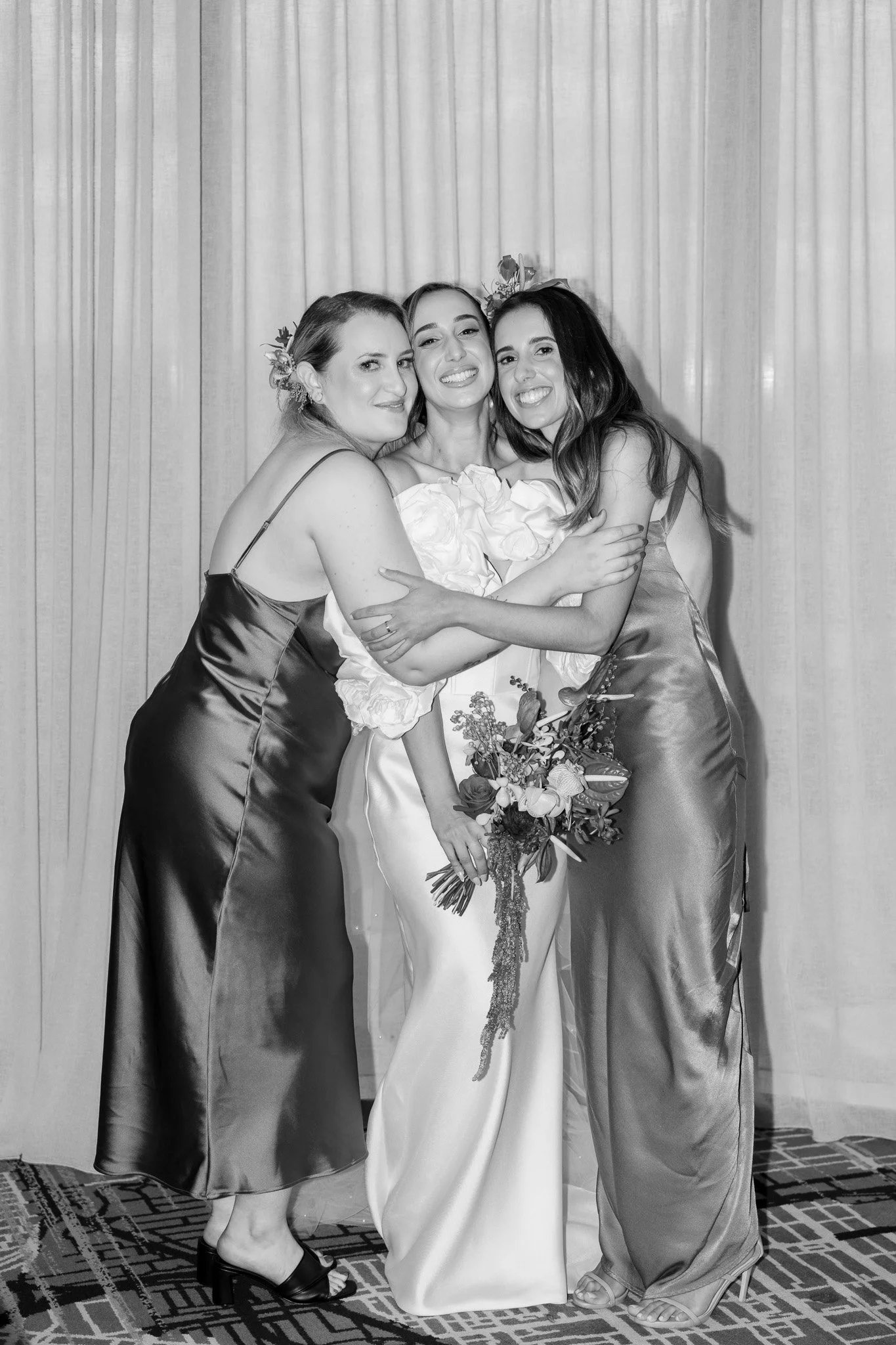 Three women in formal dresses hugging and smiling in front of a curtain, one holding a bouquet of flowers.