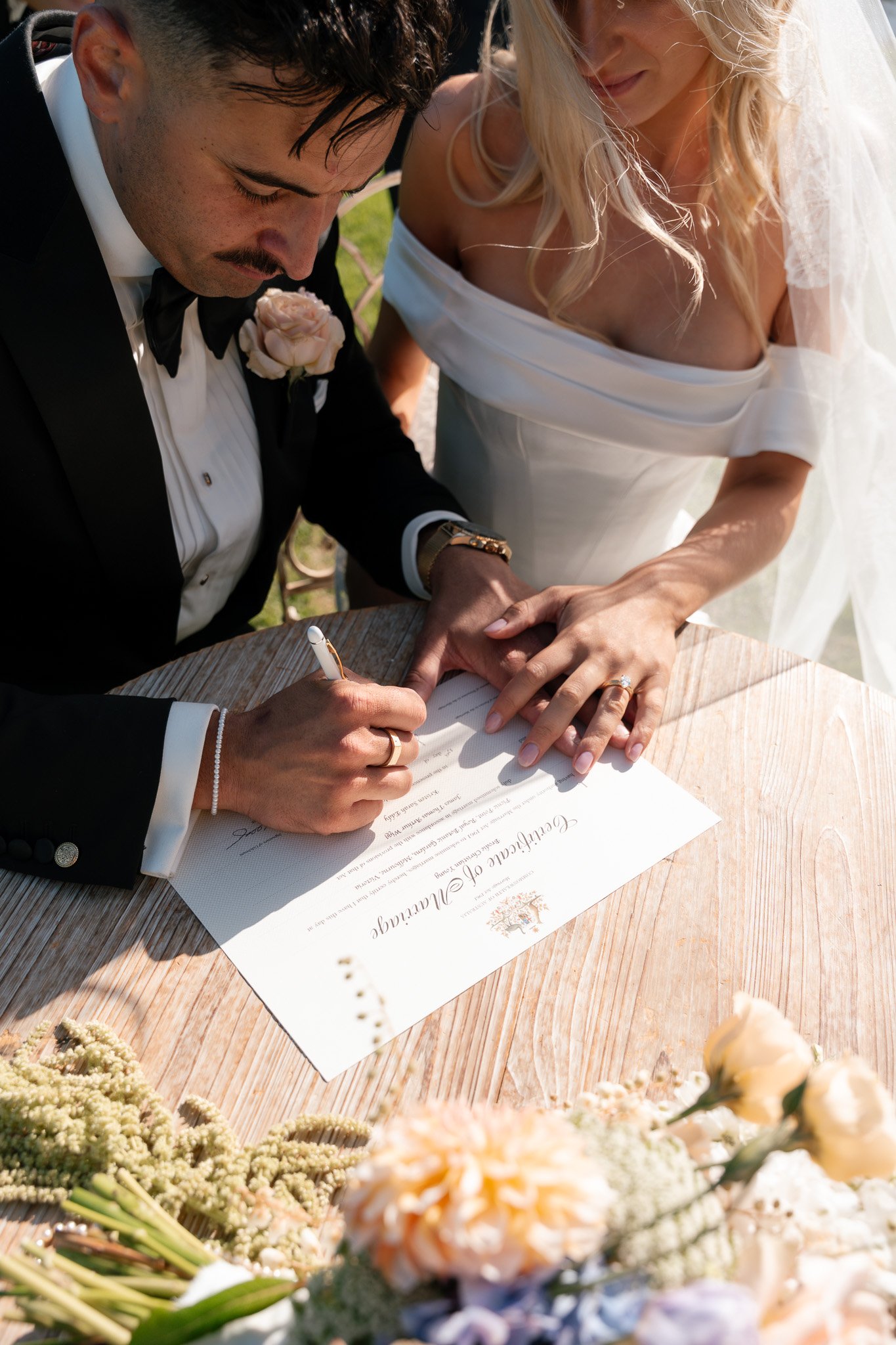 A bride and groom signing a wedding certificate outdoors, with the bride wearing a white off-shoulder dress and the groom in a black tuxedo, surrounded by flowers.