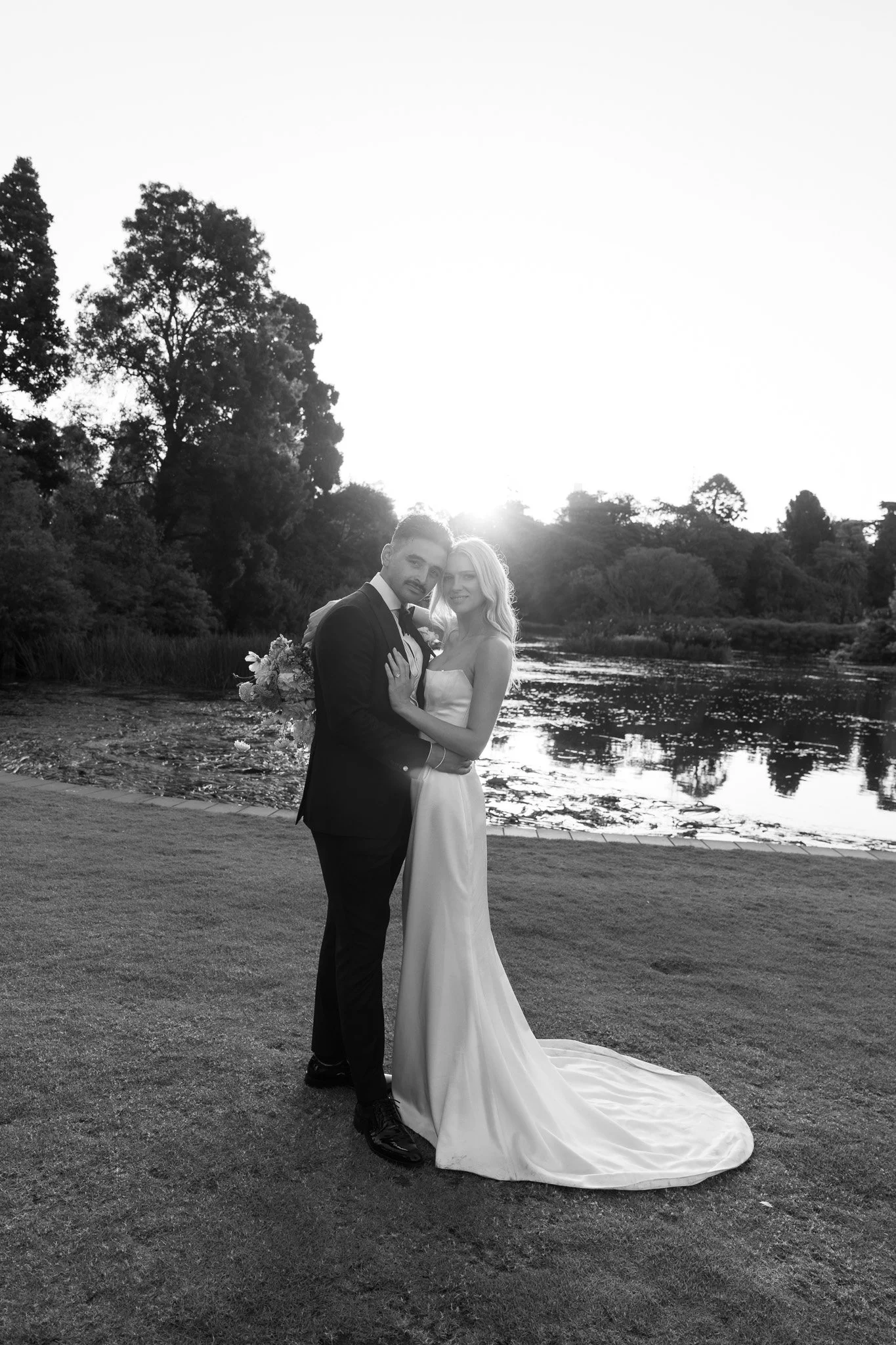 A black and white photograph of a newlywed couple embracing outdoors near a lake with trees in the background at sunset.