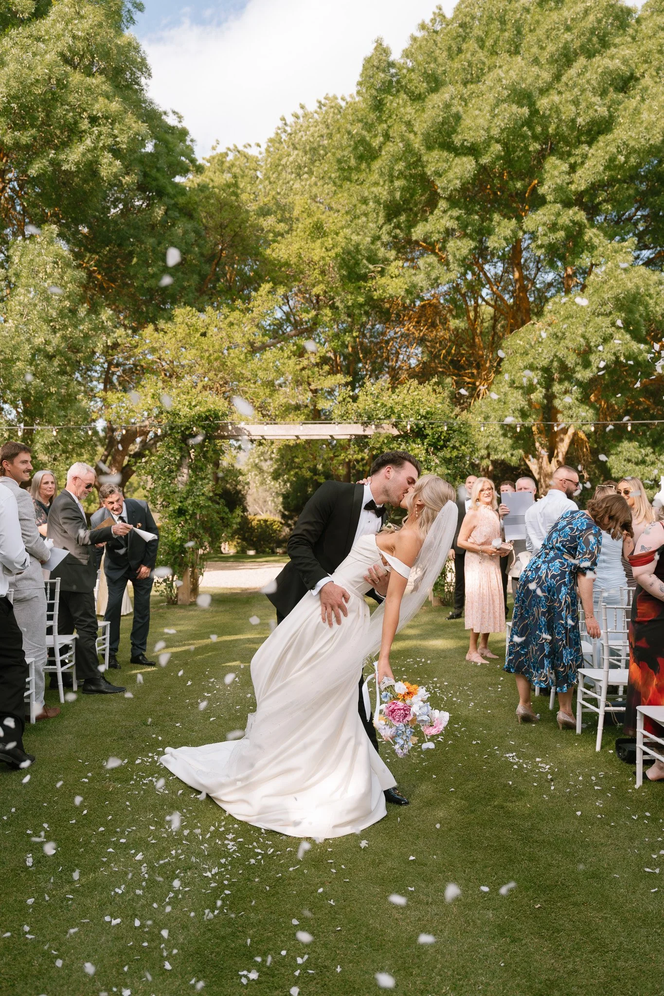 A newlywed couple sharing a kiss while the groom dips the bride during their outdoor wedding ceremony, with guests watching and throwing flower petals under green trees on a sunny day.