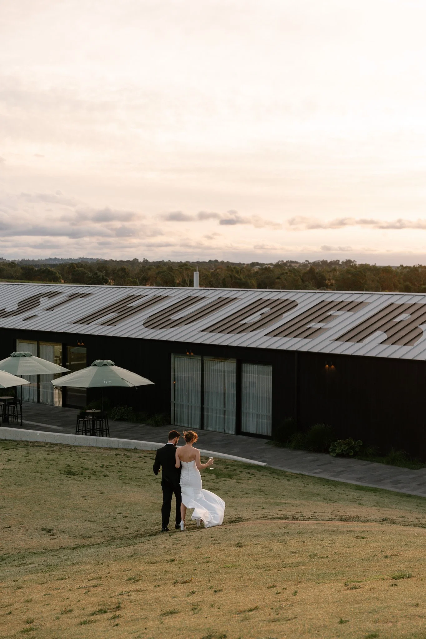 A bride and groom walking on a grassy area near a modern building with outdoor seating and umbrellas.