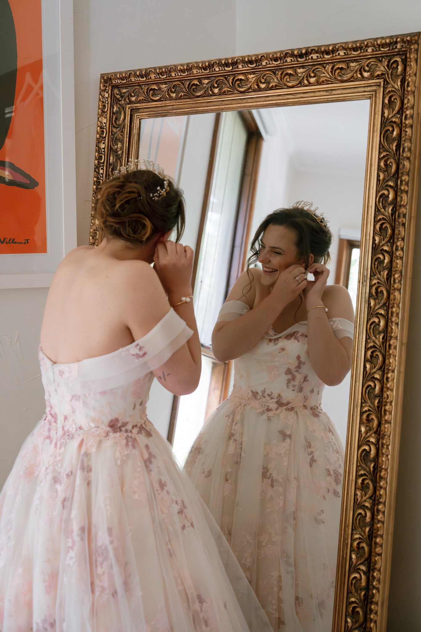 A woman in a floral dress is smiling and adjusting her earring in front of a large ornate gold mirror, with her reflection visible.