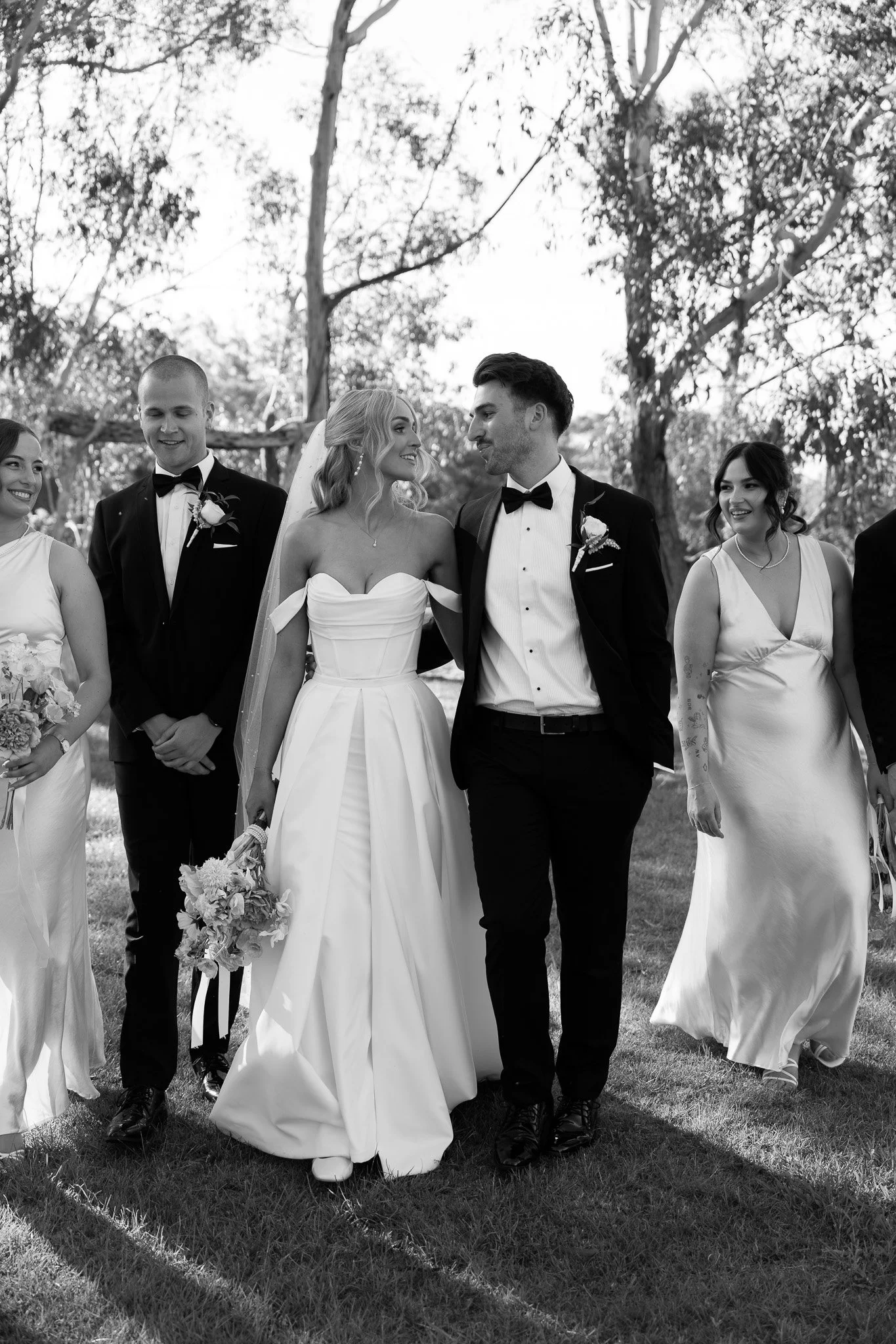 Black and white photo of a wedding procession outdoors. The bride and groom walk arm-in-arm, surrounded by bridesmaids and groomsmen, with trees in the background.