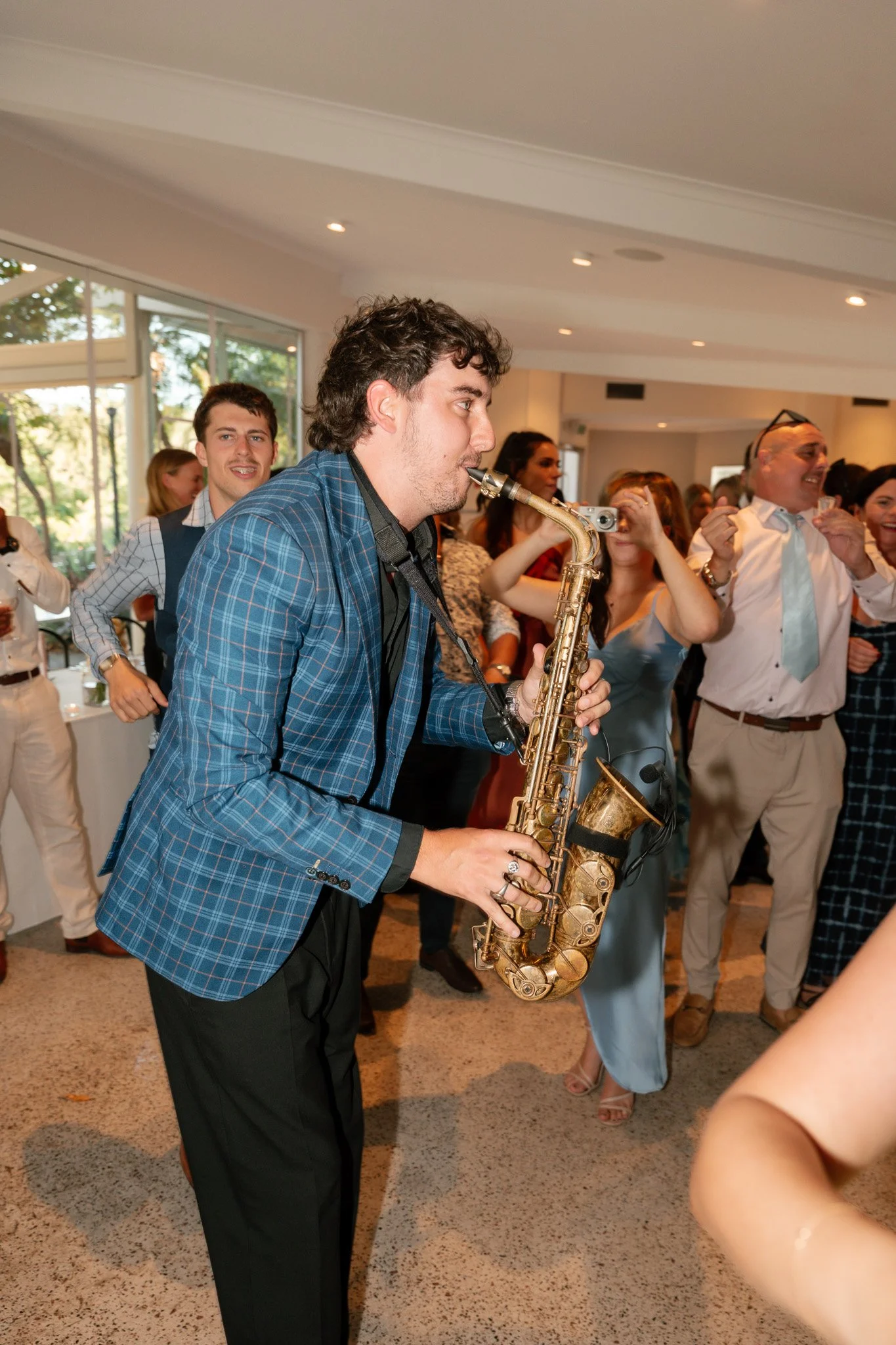 A man playing saxophone at a lively indoor event with several people dancing and enjoying the music.