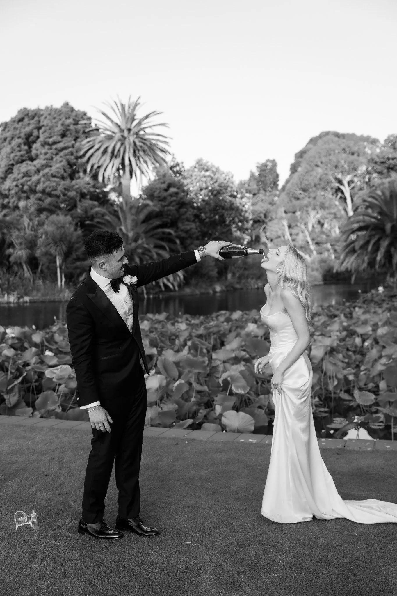 A black and white photo of a groom in a tuxedo and a bride in a wedding dress outdoors near a pond with lily pads, with trees in the background. The groom is pouring champagne into the bride's mouth.