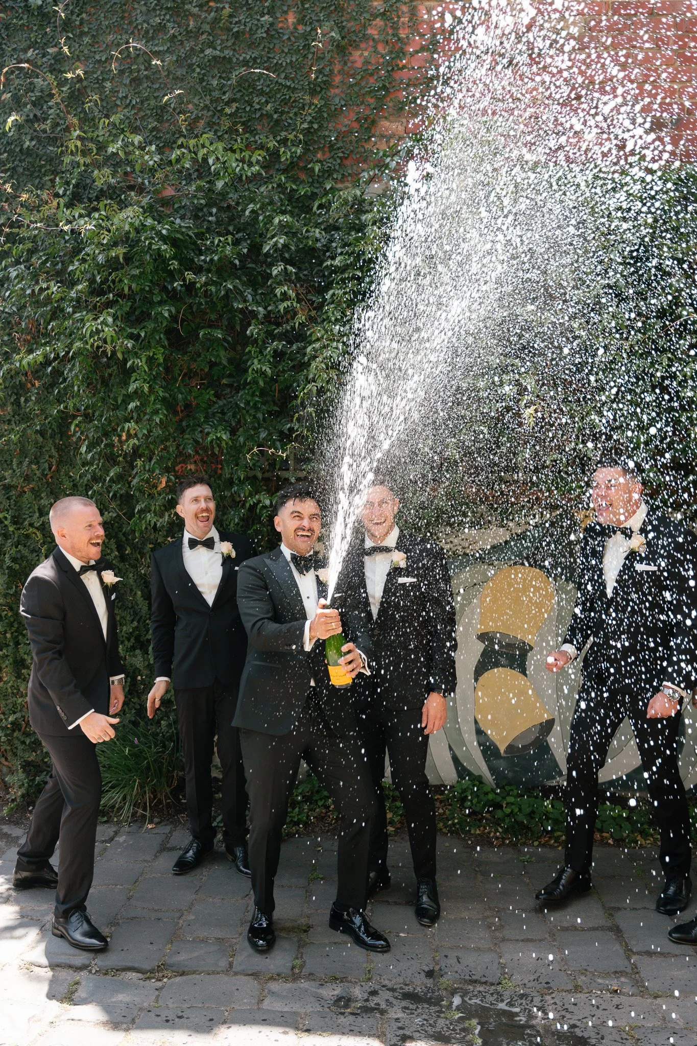 Group of men in tuxedos celebrating with a champagne cork pop and spray, outdoors on a sidewalk, with greenery and a mural in the background.