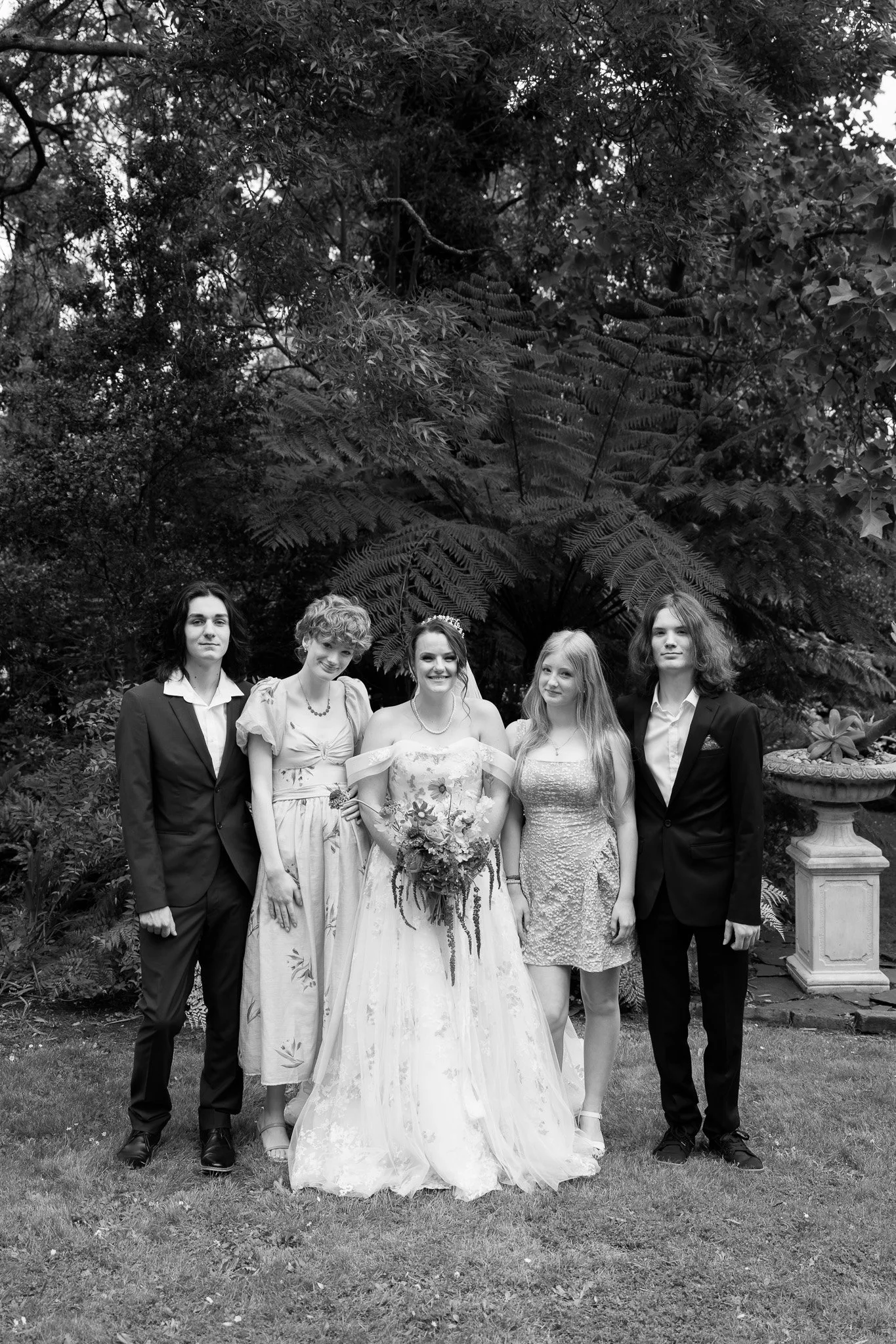 Five young women at a wedding in a garden, with one in a wedding dress holding a bouquet, and the others in formal dresses and suits, standing in front of lush trees and plants.