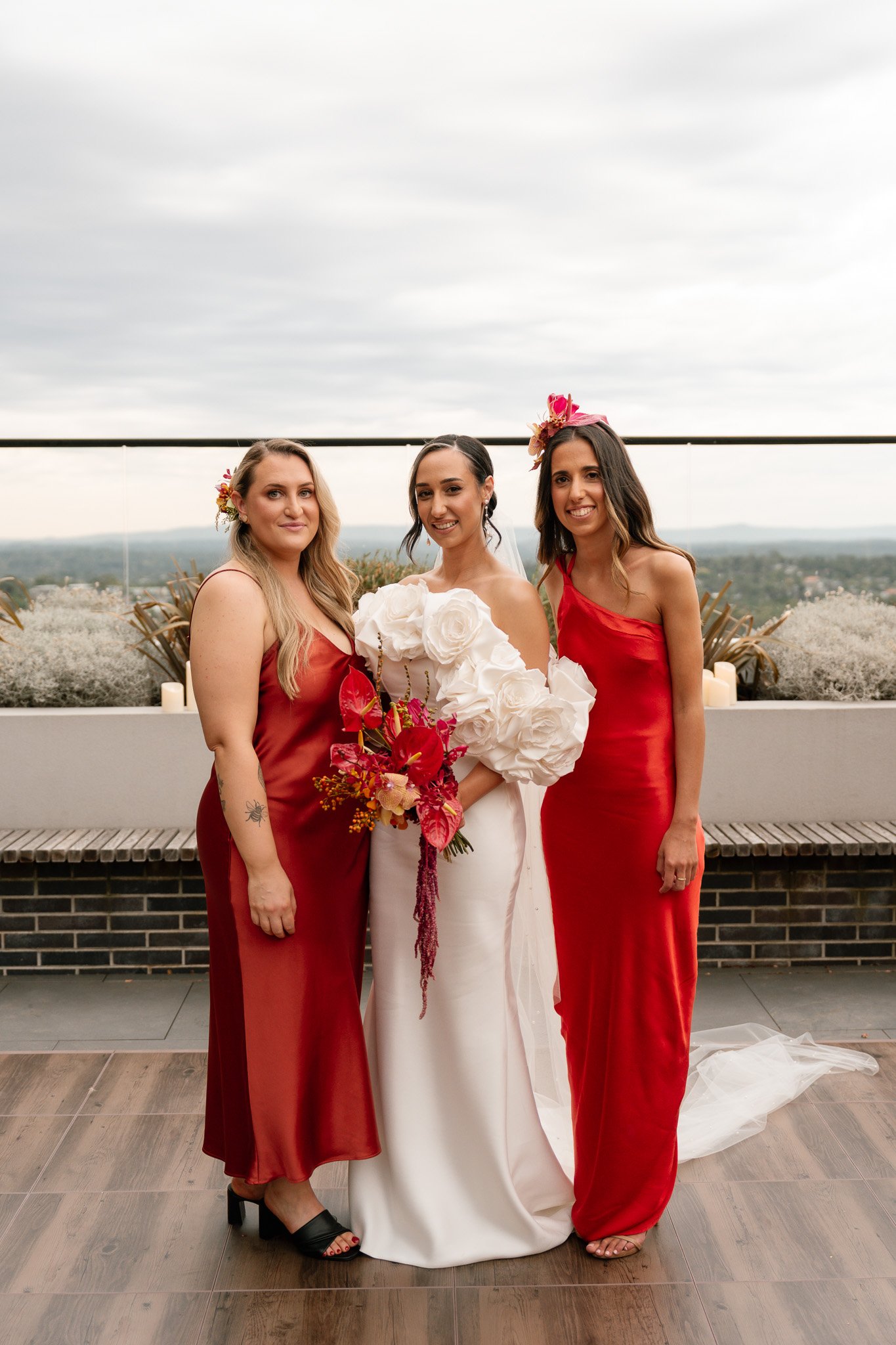 Three women in evening gowns, one in a white wedding dress holding a bouquet, standing outdoors with cloudy sky and landscape in the background.