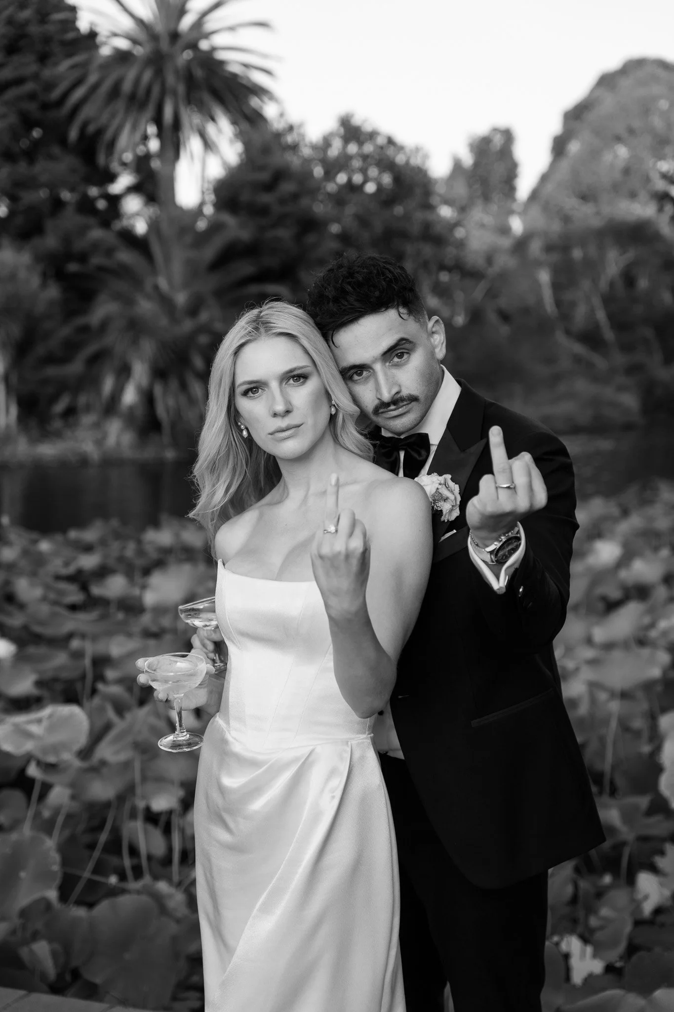 A black and white photo of a couple, a woman in a white dress and a man in a tuxedo, standing outdoors with trees and water in the background, both making rude gestures with their middle fingers.