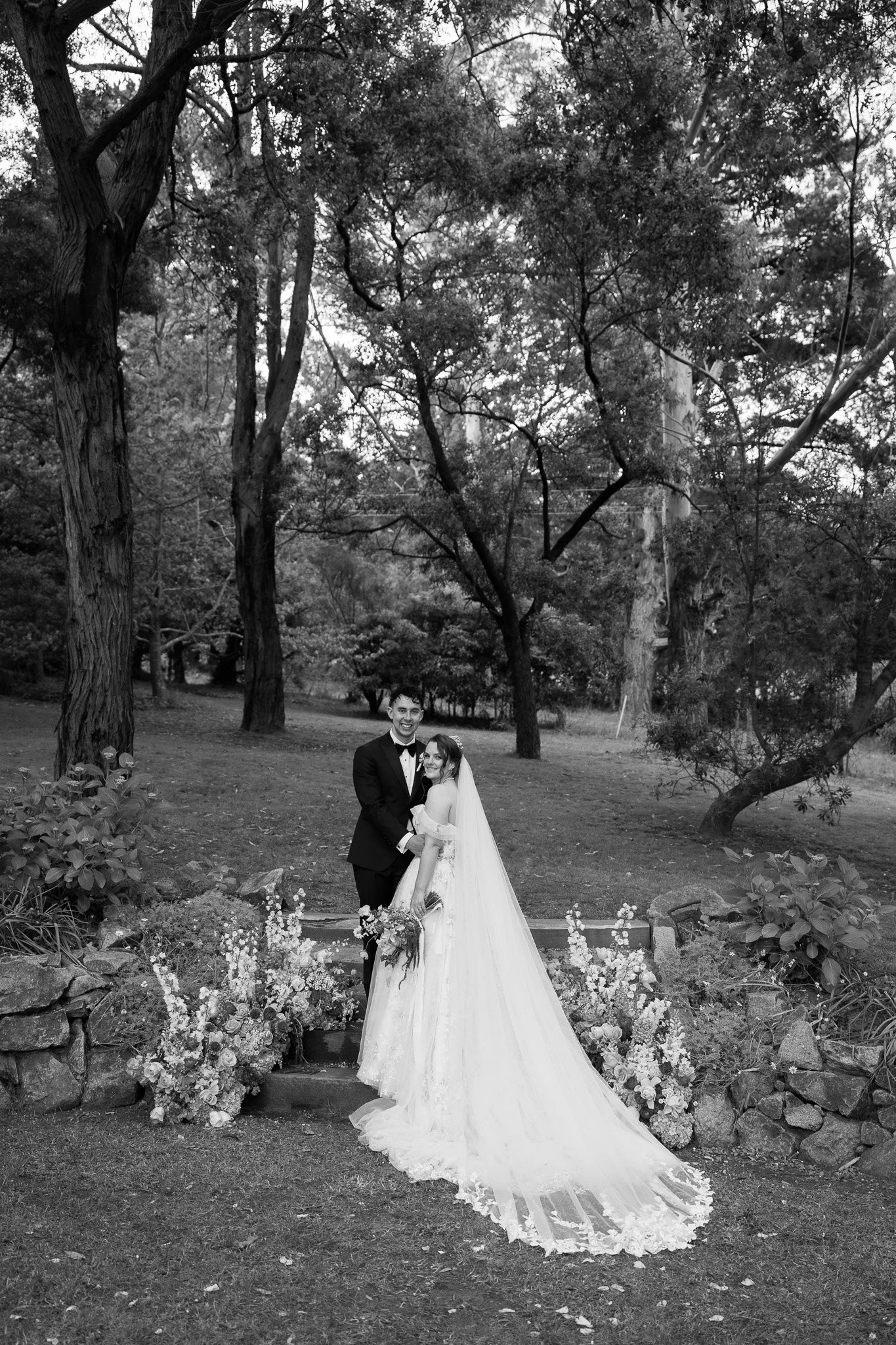 Black and white photo of a newlywed couple standing outdoors on a stone platform surrounded by flowers and trees; the bride wears a long wedding gown with a veil, and the groom is in a tuxedo.