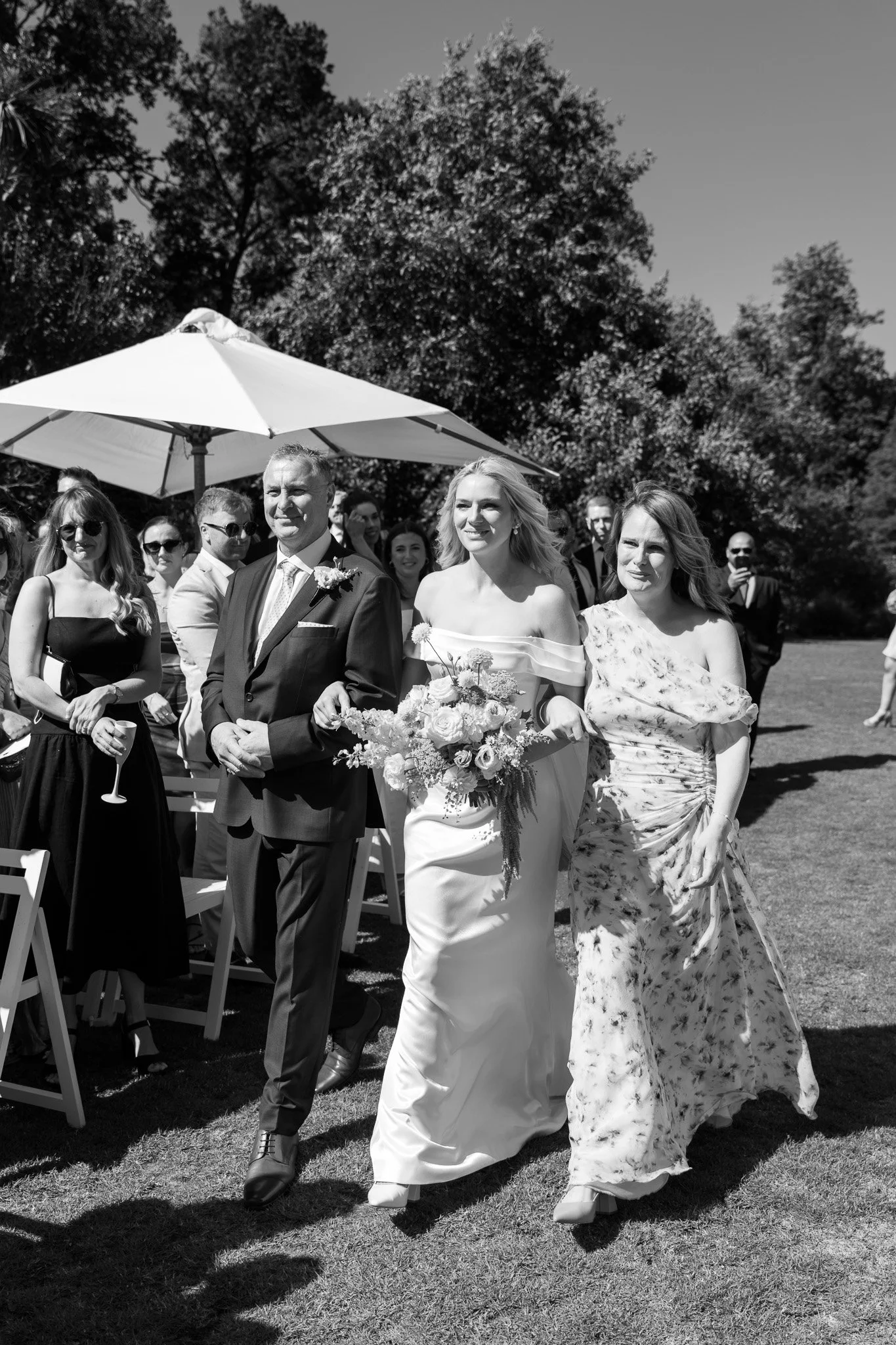 A bride walking down the aisle with her father at an outdoor wedding ceremony, surrounded by guests seated and standing under a parasol on a sunny day.