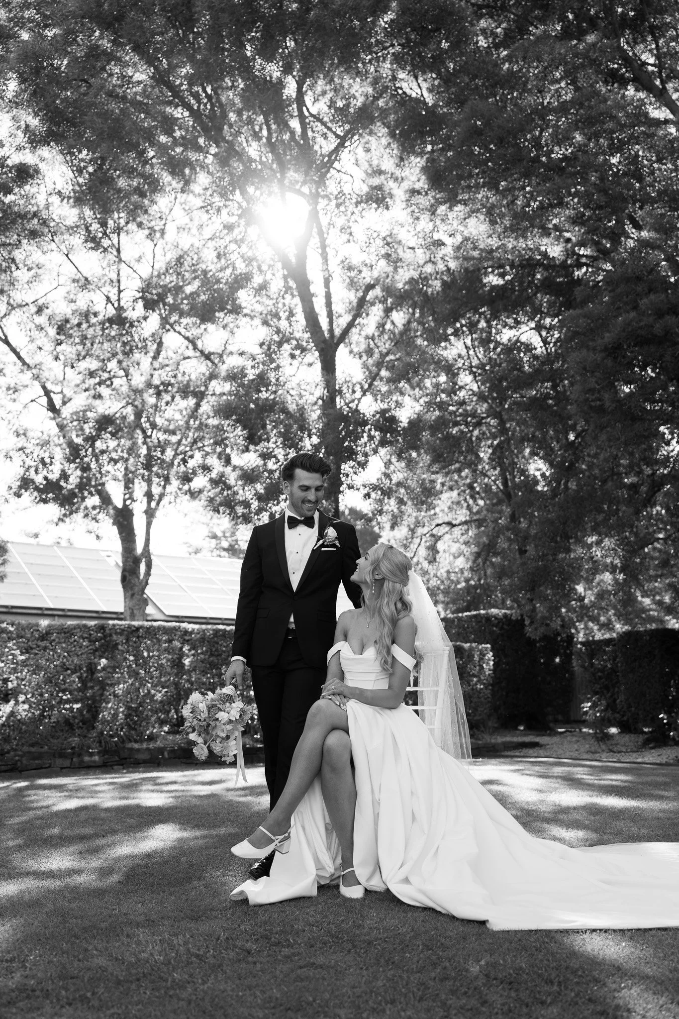 Black and white photo of a wedding couple outdoors; the groom in a tuxedo, the bride in a wedding dress, sitting on the grass, the groom holding a bouquet, with trees and sunlight in the background.