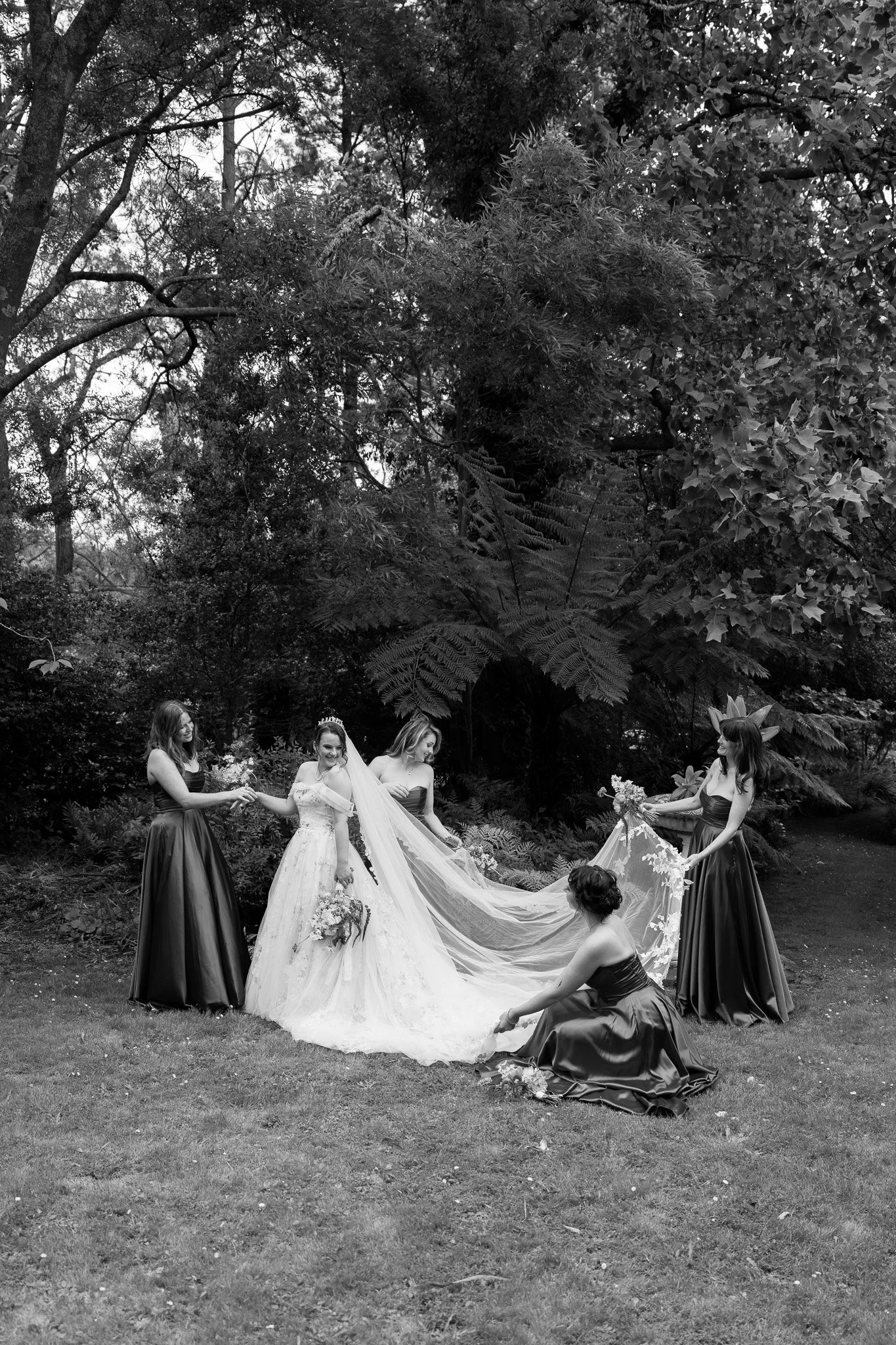A black-and-white photo of a bride and four bridesmaids outdoors among trees, with the bride holding a bouquet and the bridesmaids assisting her with her long veil.