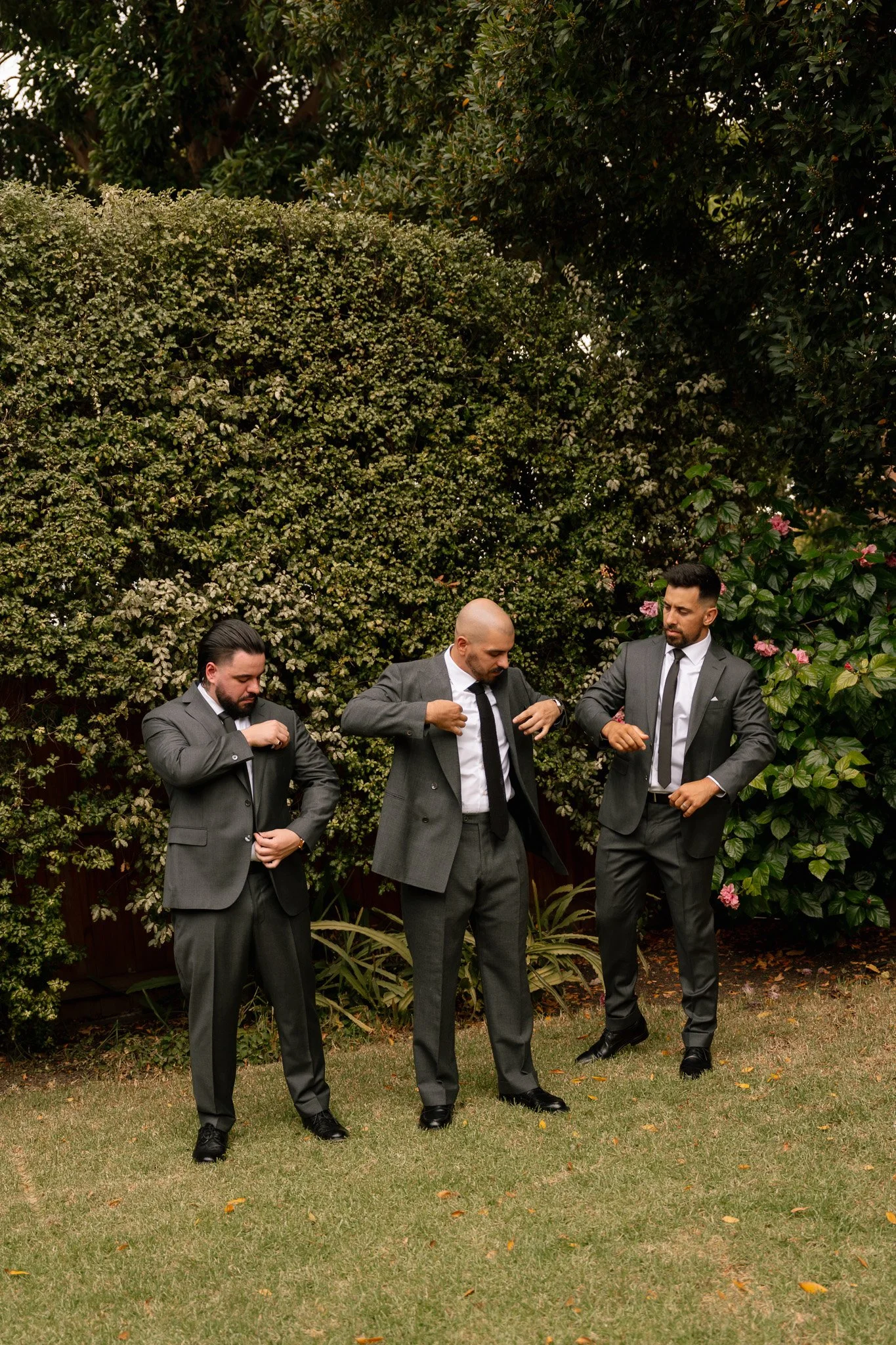 Three men in suits adjusting their ties outdoors in a garden with bushes and flowers.