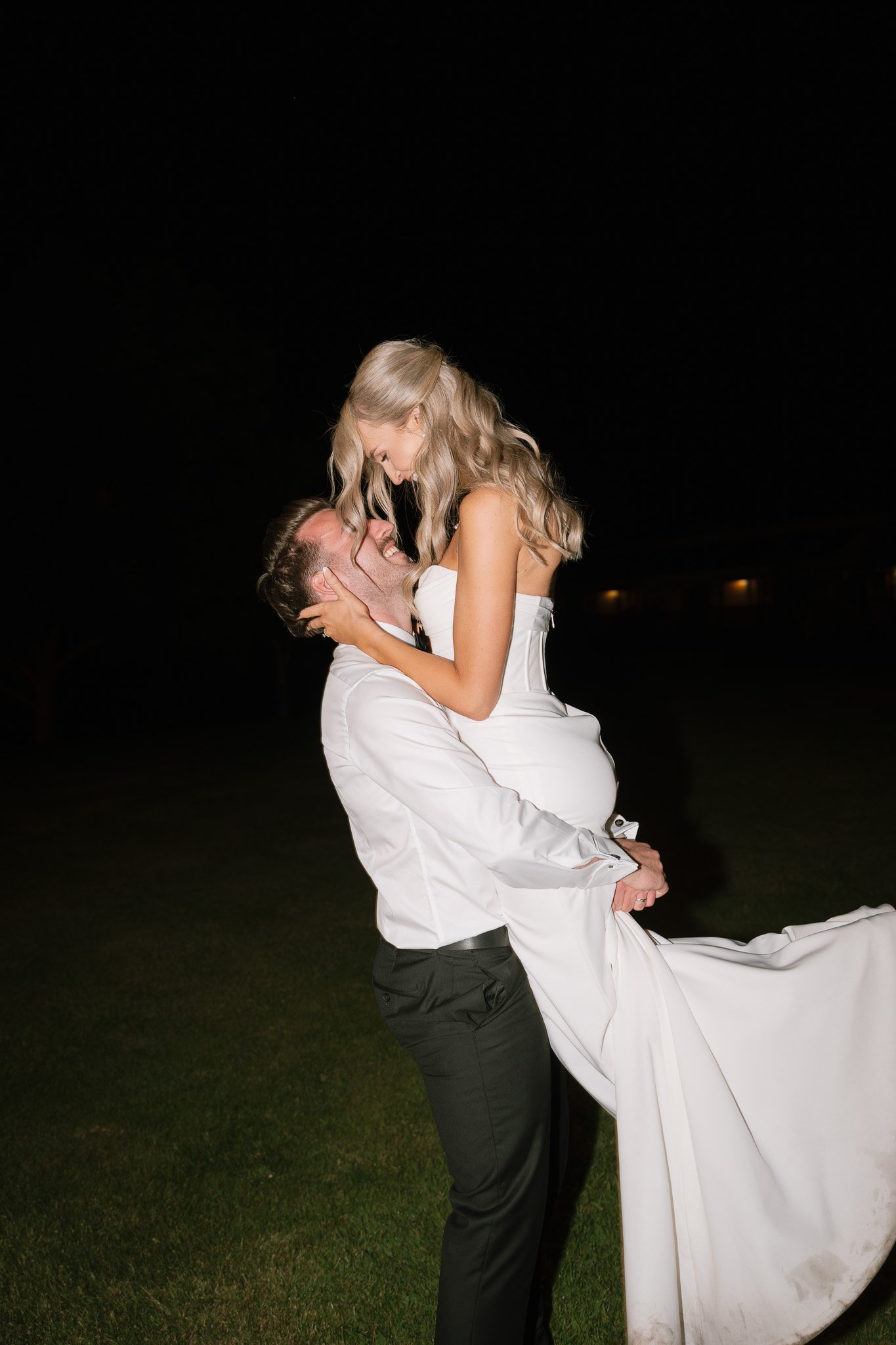 A man in black pants and a white shirt lifting a woman in a white wedding dress into the air during nighttime, all smiling and looking happy.