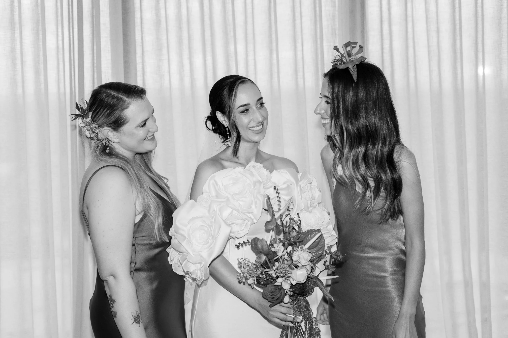 Three women at a wedding, with the bride in the center holding a bouquet of flowers, flanked by two women in dresses, all smiling at each other, with a curtain backdrop.