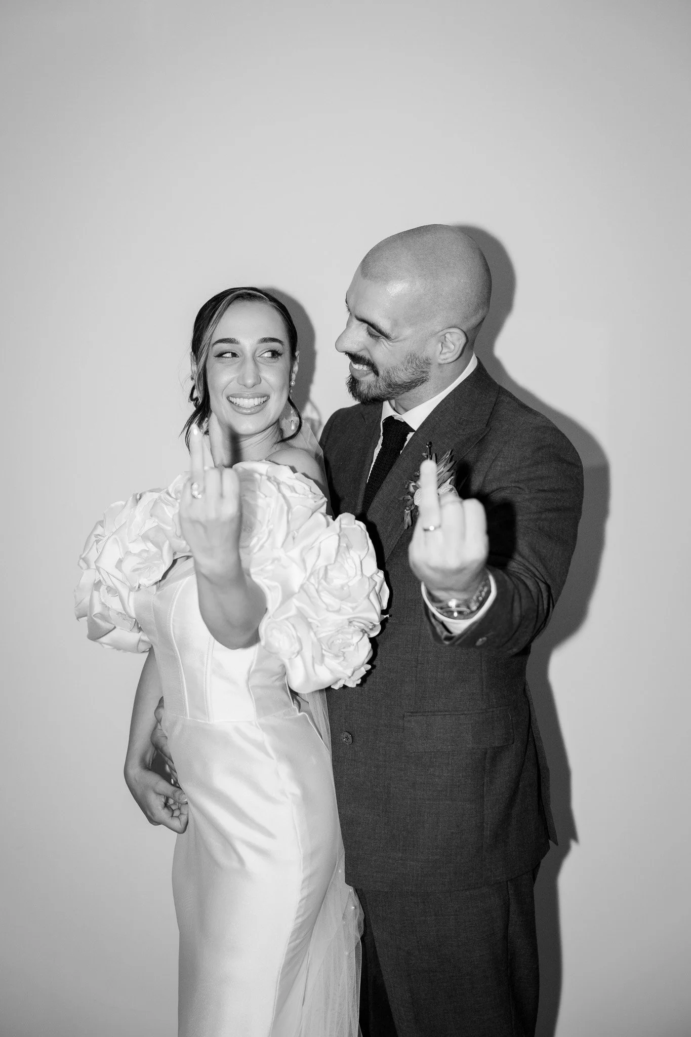 Black and white photo of a bride and groom smiling and showing their middle fingers.