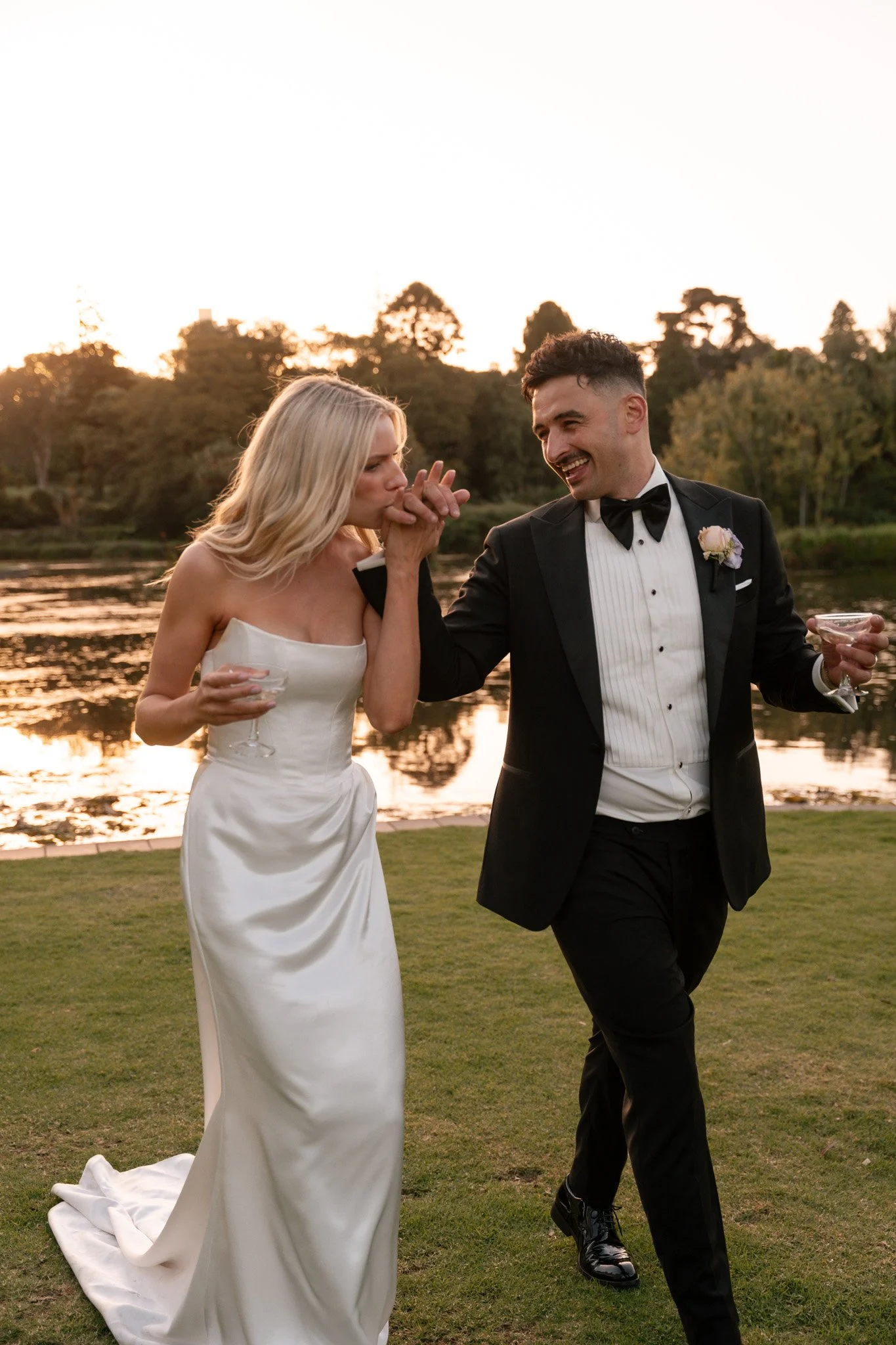 Couple dressed in wedding attire enjoying a moment by a lake during sunset. The bride is in a white gown, and the groom is in a black tuxedo. They are smiling and holding drinks.