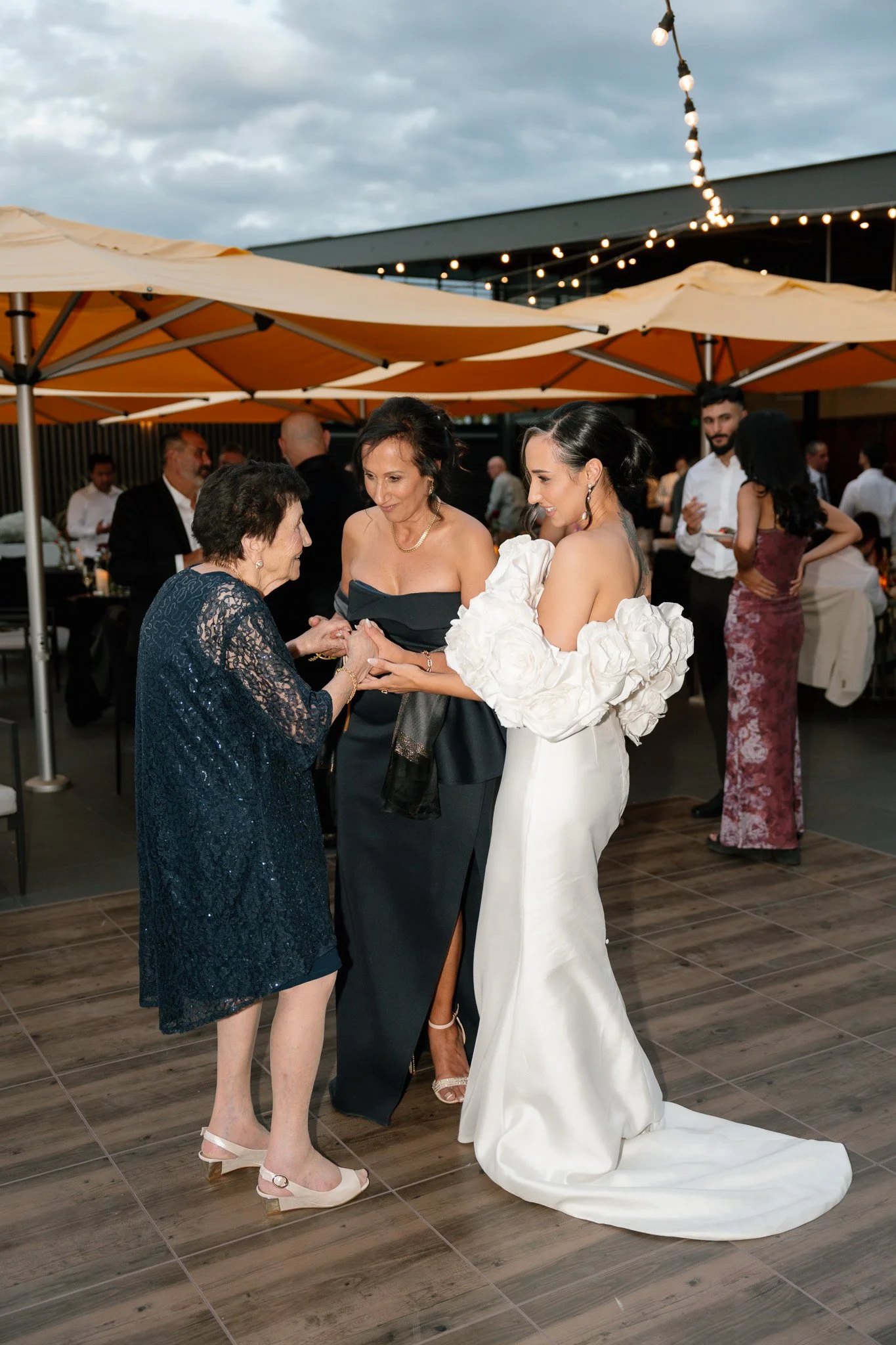 Three women at a wedding reception, holding hands and smiling, with other guests in the background under patio umbrellas and string lights.