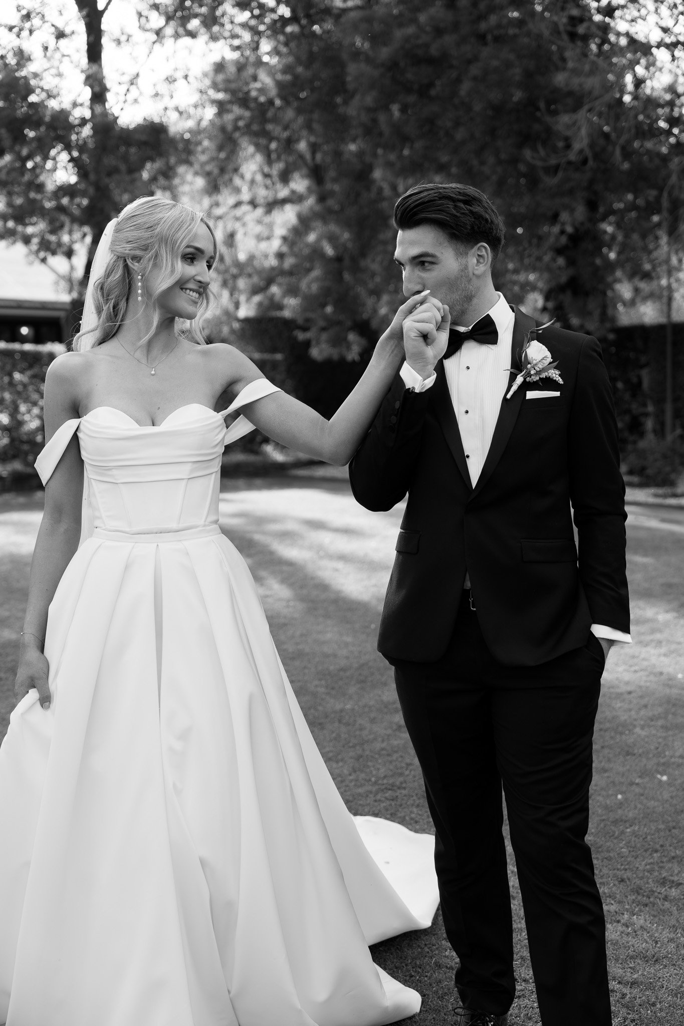A black and white photo of a bride and groom outdoors, with the bride gently touching the groom's nose. The bride is smiling, wearing a strapless wedding dress, and the groom is wearing a tuxedo with a bow tie. Trees and a lawn are in the background.
