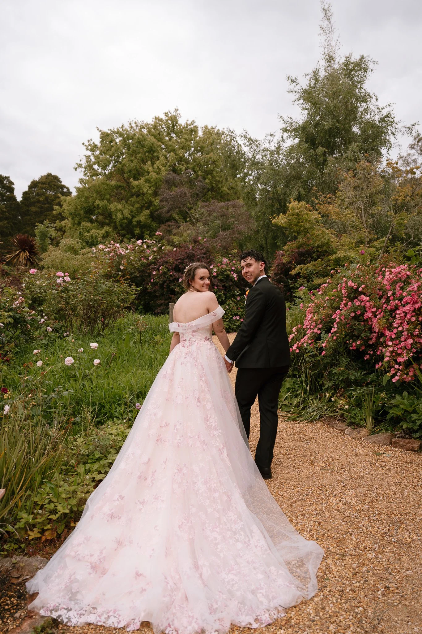 A bride in a pink wedding gown and a groom in a black suit holding hands and walking down a garden path surrounded by blooming pink and purple flowers and green trees.