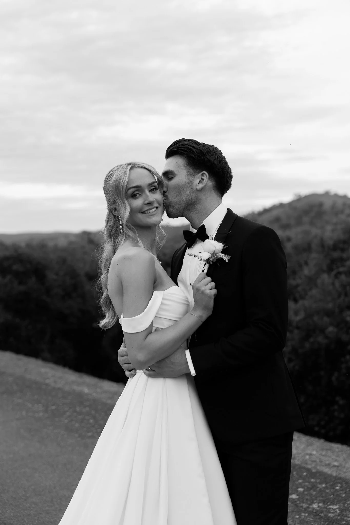 Black and white photo of a bride and groom on their wedding day. The bride is smiling and holding a boutonniere, while the groom is kissing her on the temple. Hills and cloudy sky in the background.