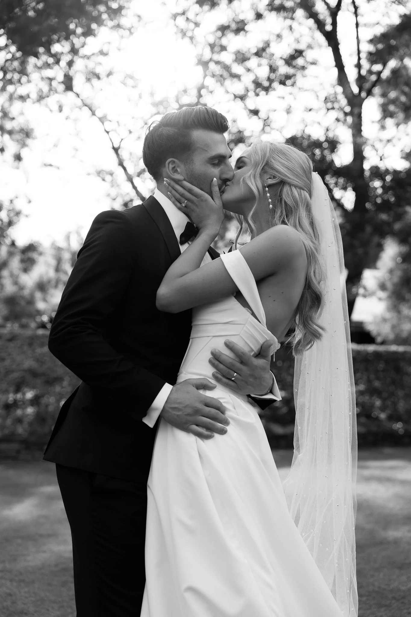 A black-and-white photo of a bride and groom kissing outdoors, with trees in the background. The bride has long wavy hair, wears a wedding dress with a fitted bodice and off-the-shoulder sleeves, and a veil. The groom has short, styled hair, wears a 