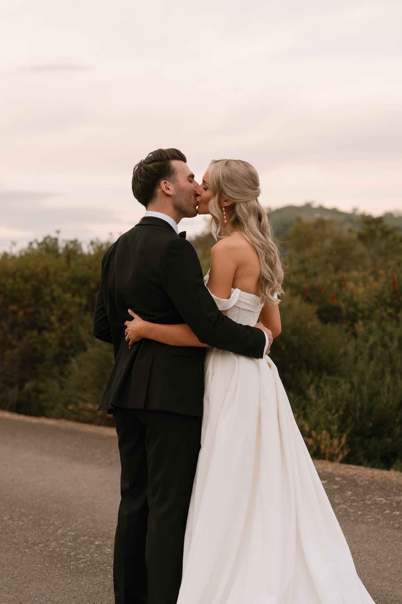 A newlywed couple sharing a kiss outdoors at sunset, with the bride in a white off-the-shoulder gown and the groom in a black tuxedo, embracing on a paved path with greenery and trees in the background.