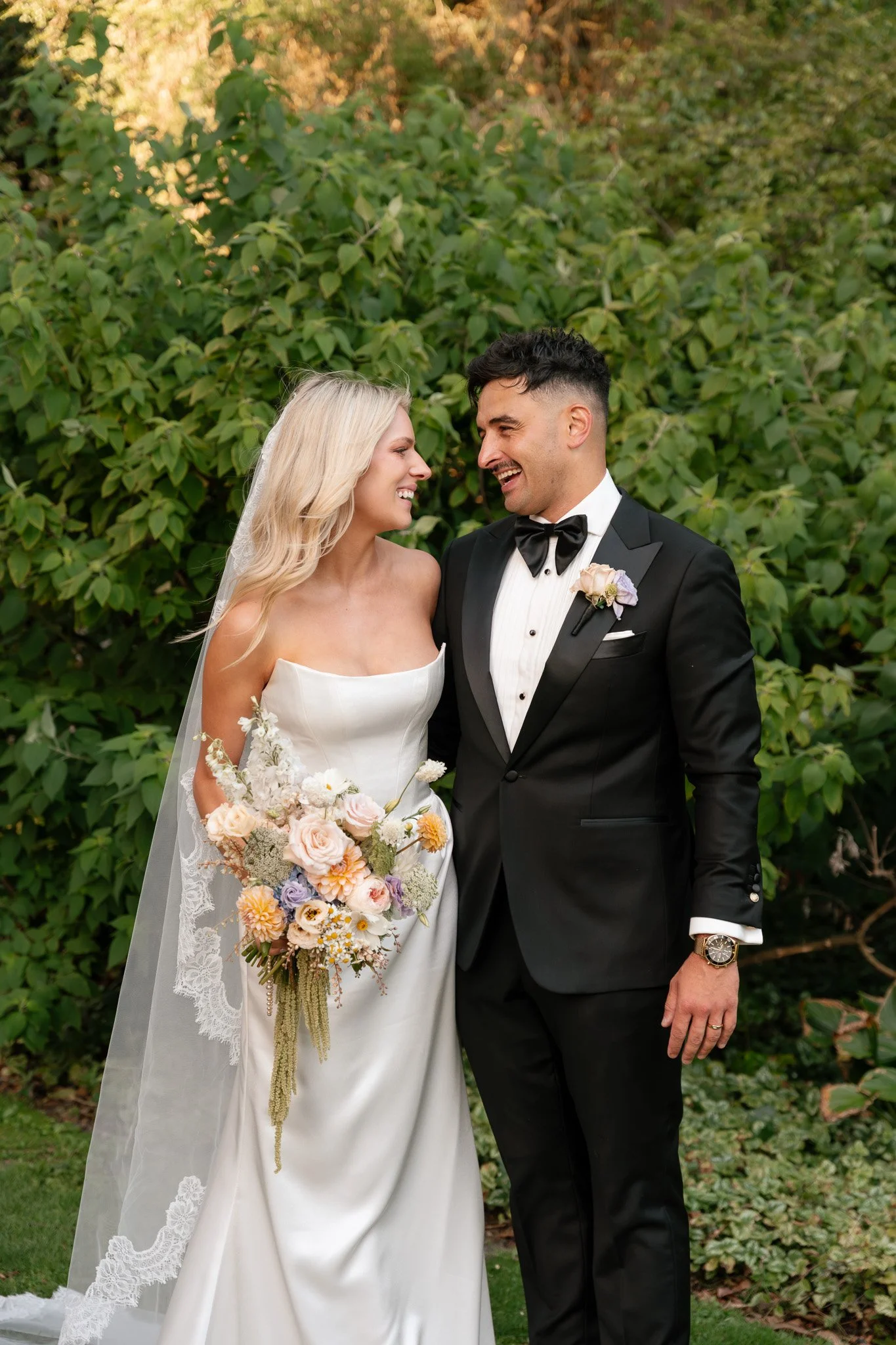 A bride and groom smiling and looking at each other outdoors, with a green leafy background. The bride is wearing a strapless white wedding gown and veil, holding a bouquet of pastel-colored flowers. The groom is dressed in a black tuxedo with a bow 