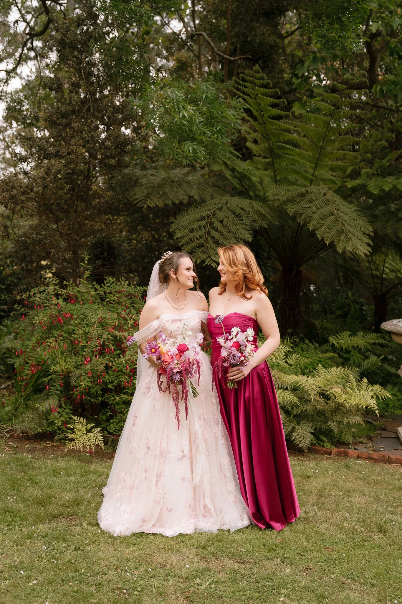 Two women in wedding attire, standing outdoors, smiling and holding bouquets of pink and purple flowers, surrounded by lush greenery.