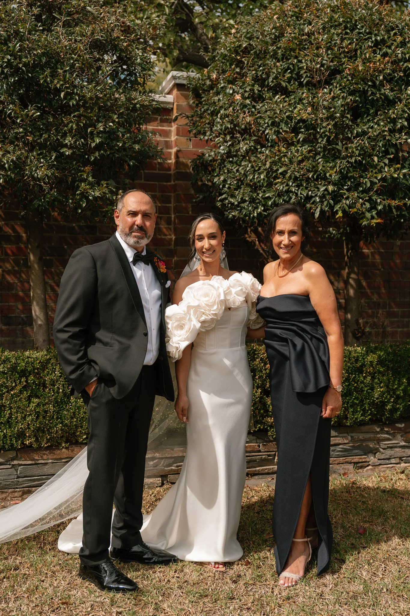 Three people standing outdoors in front of bushes and a brick wall, dressed formally for a wedding, including a bride in a white gown with large floral embellishments on the sleeves, a man in a tuxedo, and a woman in a strapless black gown.