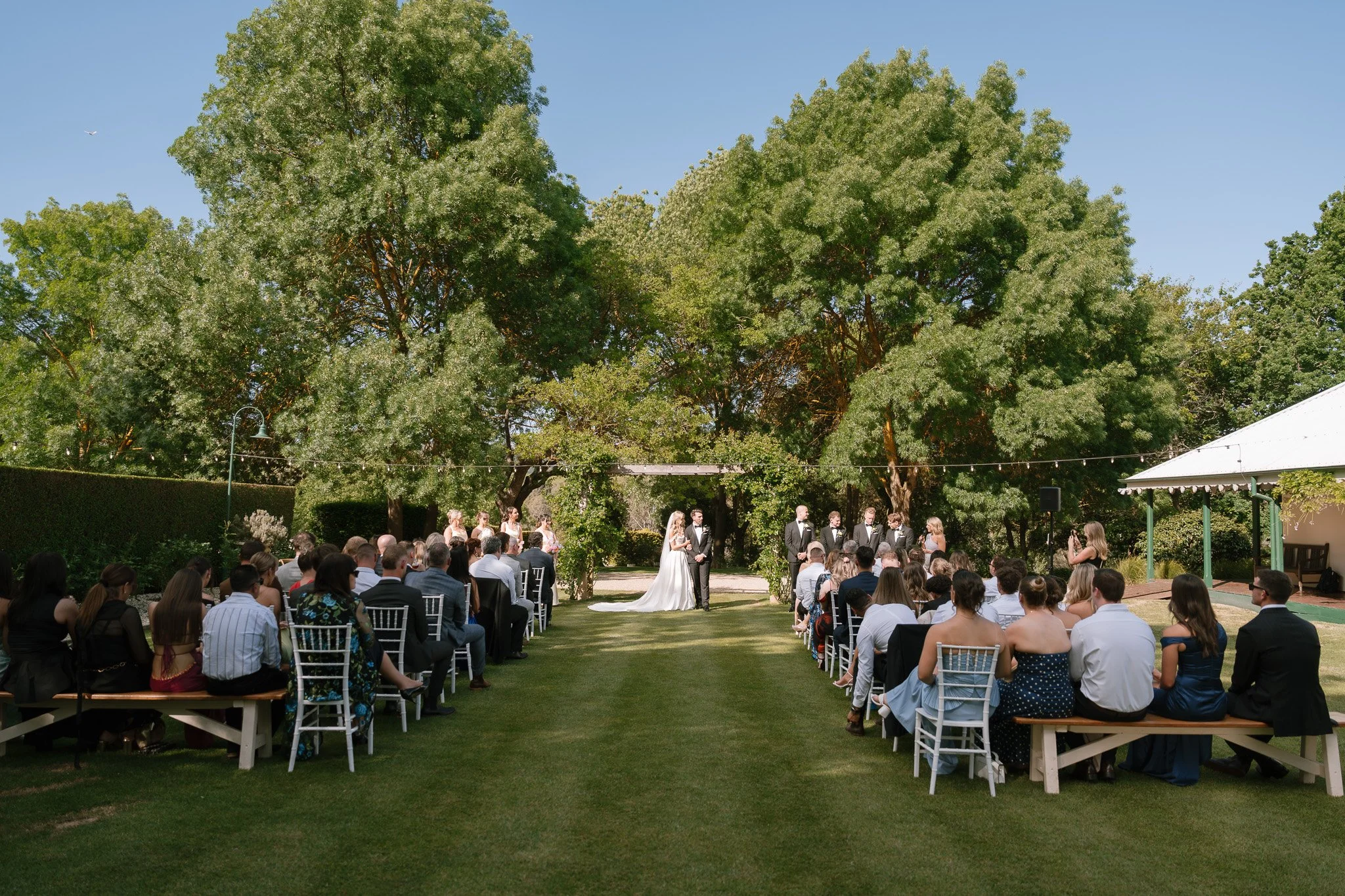 Outdoor wedding ceremony with bride and groom standing under a large tree, surrounded by seated guests, during daytime with clear sky.