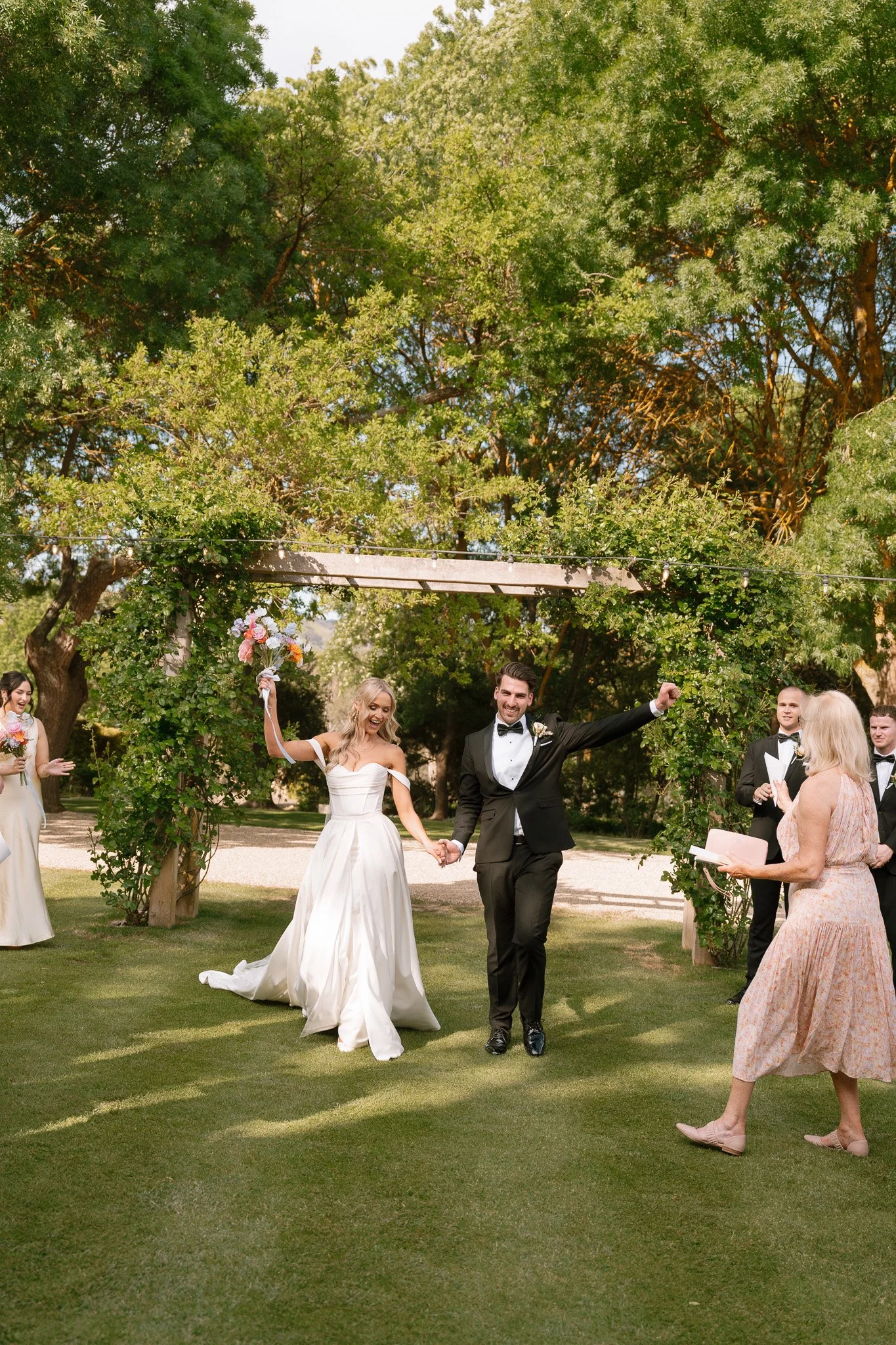 Wedding ceremony outdoors with bride and groom holding hands, bride holding a bouquet, and guests applauding under a decorated arch and green trees.