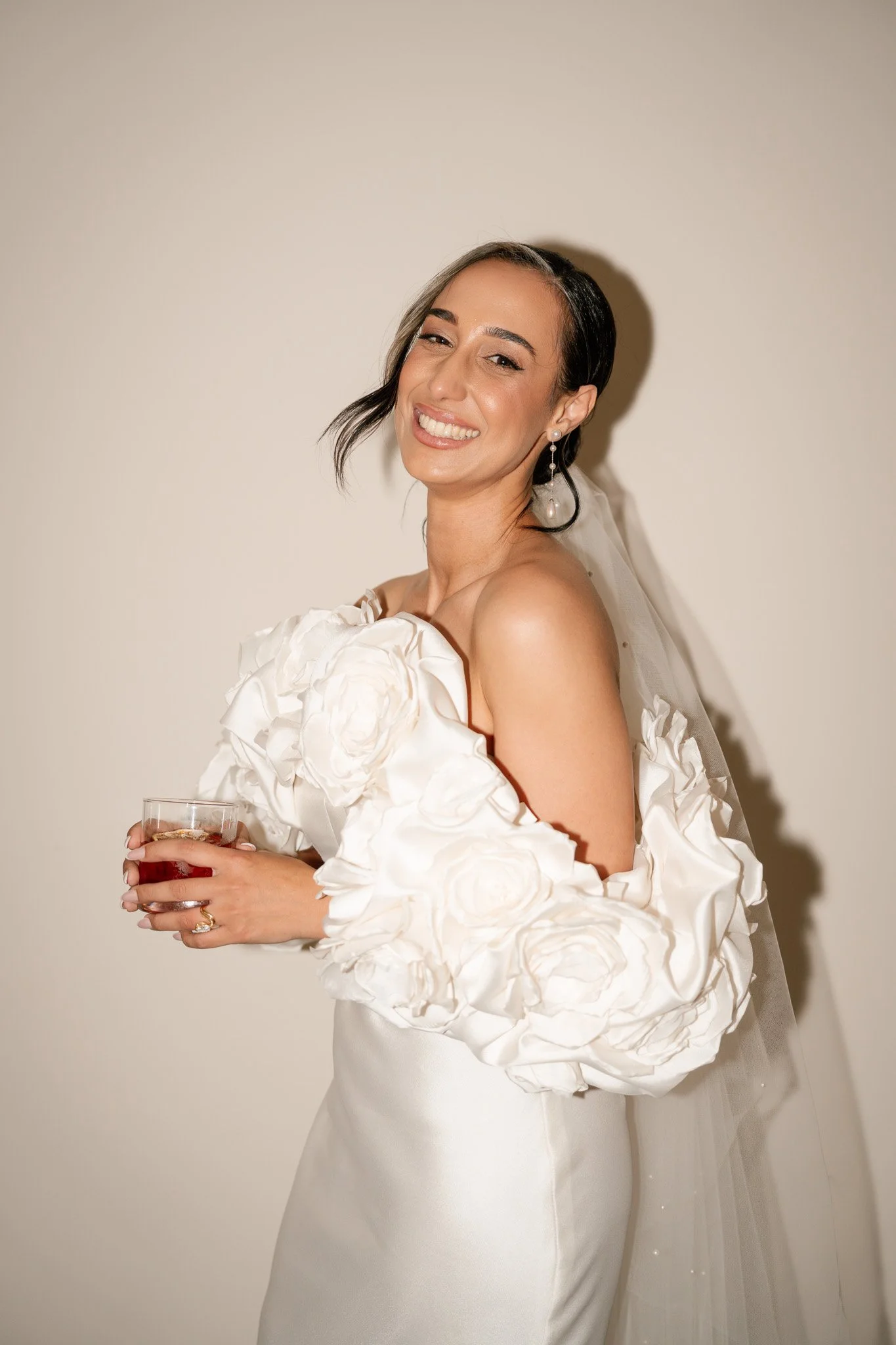 A woman in a white wedding dress with large floral fabric details on the sleeves, holding a glass and smiling.