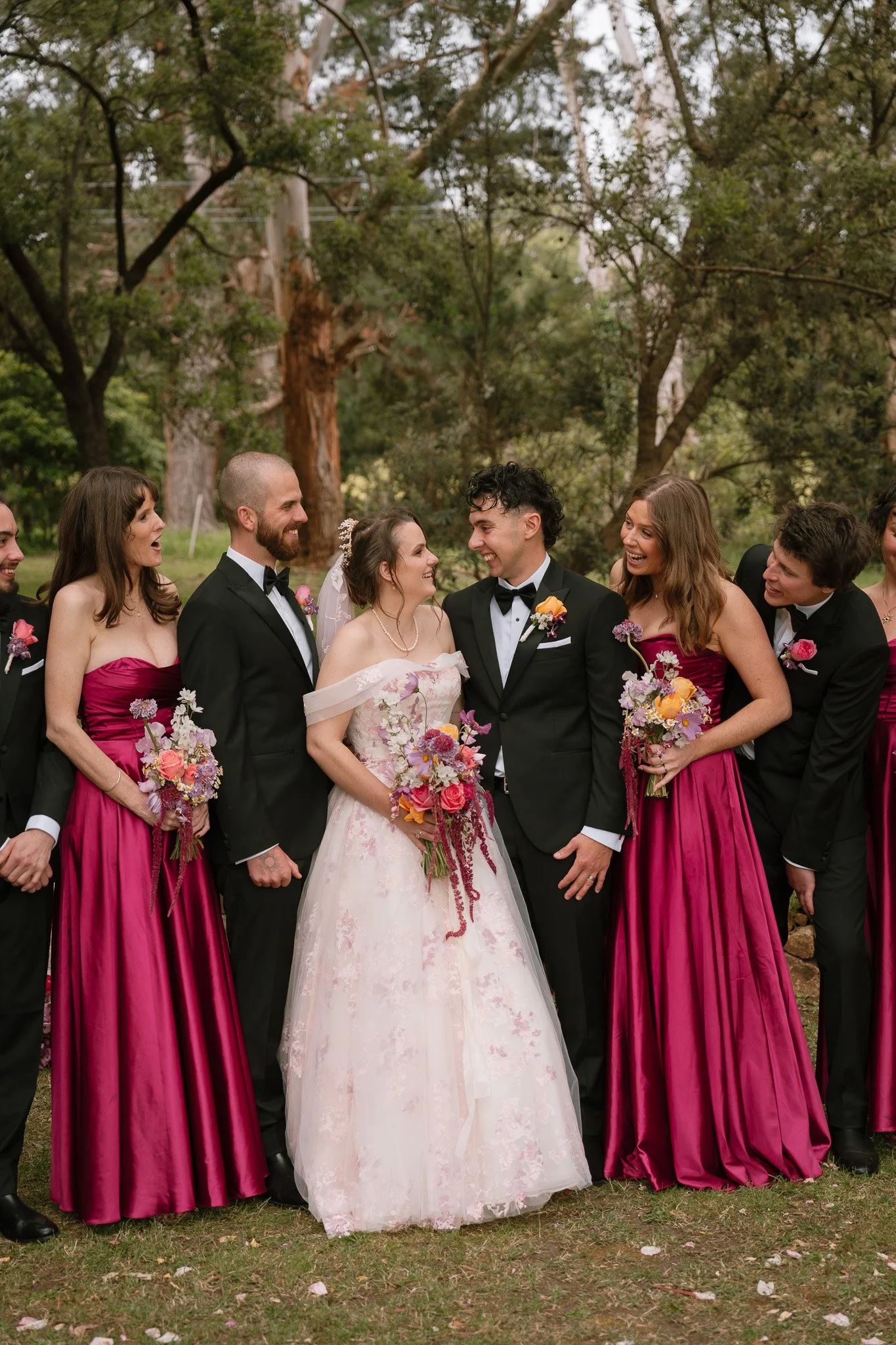 A wedding party outdoors with the bride and groom in the center, surrounded by bridesmaids in magenta dresses and groomsmen in black suits, all smiling and holding bouquets of flowers.