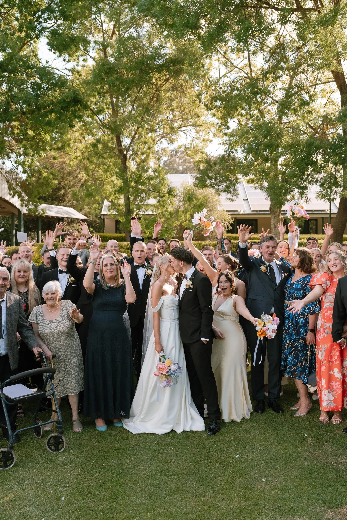 A group of wedding guests celebrating outdoors, with a bride and groom kissing in the center. The guests are raising their hands and smiling, with some holding bouquets of flowers. The scene is set under large green trees with sunlight filtering thro