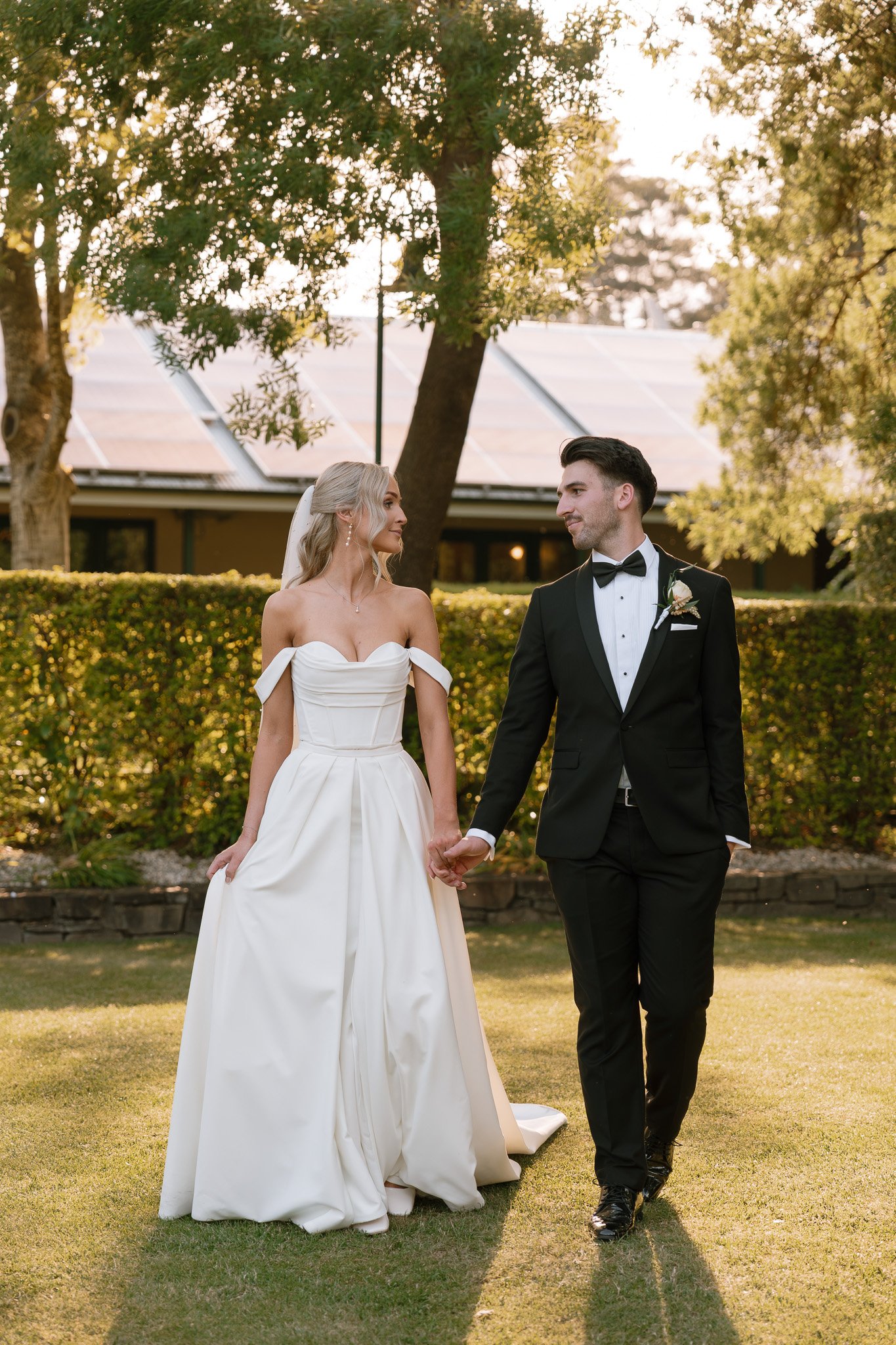 Bride and groom holding hands, walking outdoors in a garden during sunset, the bride in a white wedding gown and veil, the groom in a black tuxedo with a bow tie, surrounded by trees and a hedge.