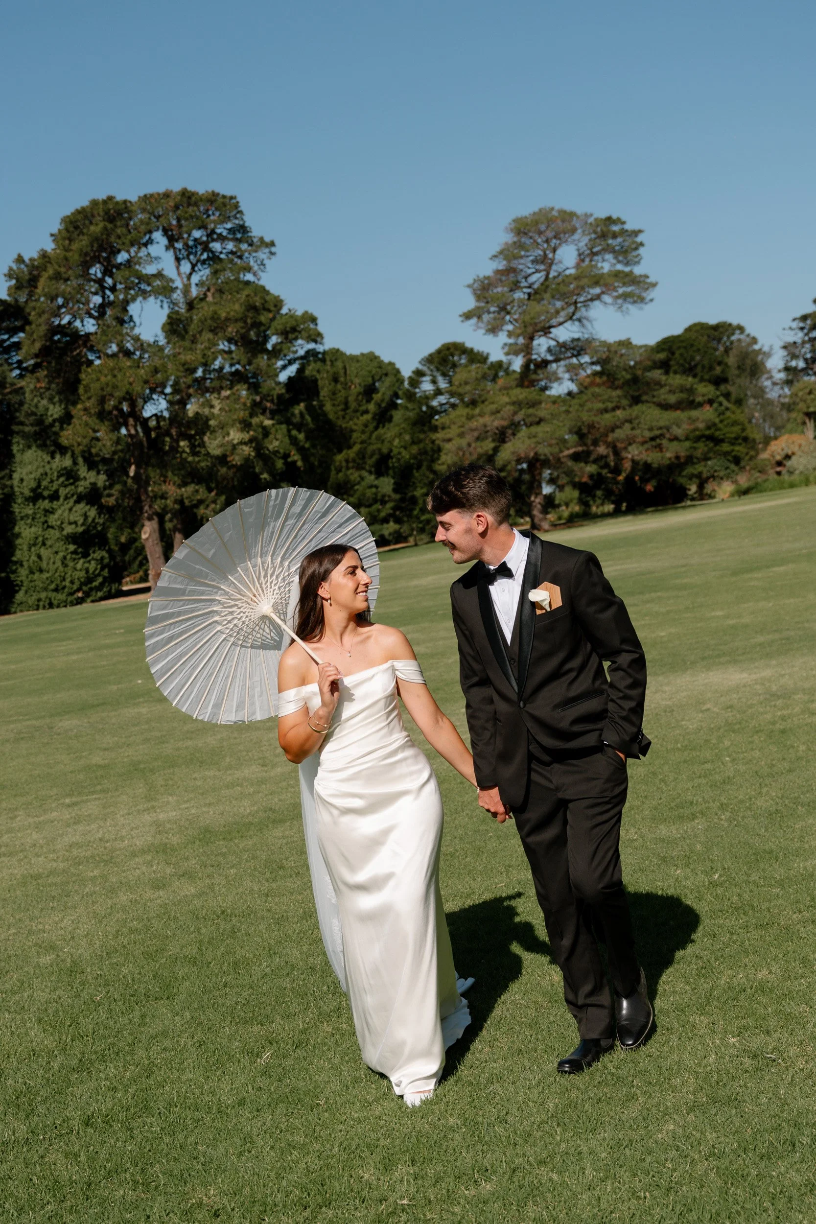 Bride and groom walking hand in hand on green lawn, bride holding a parasol, wearing white off-shoulder wedding dress, groom in black tuxedo with bow tie, trees and blue sky in background.