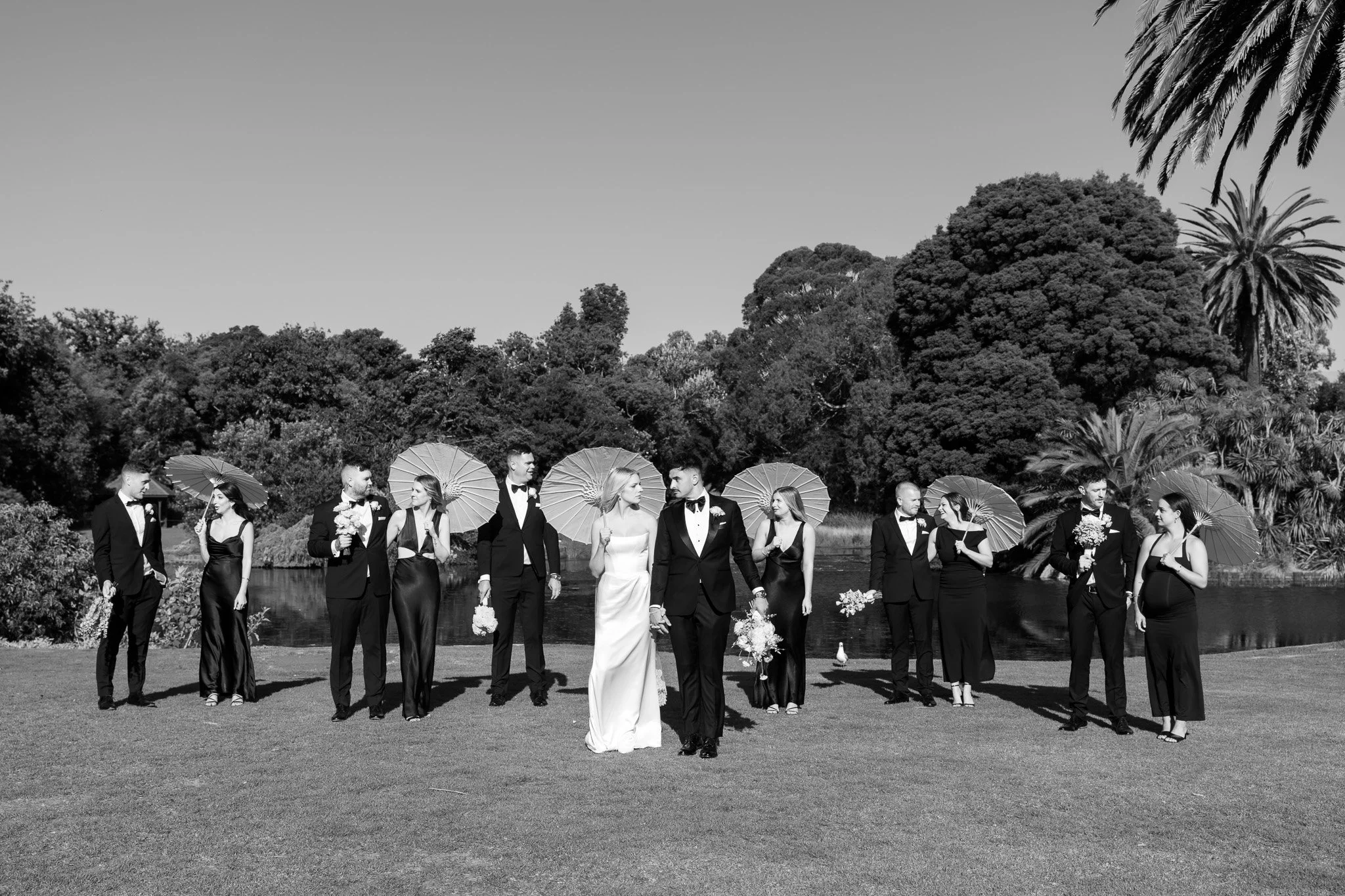 Black and white photograph of a wedding party outdoors near a lake, with the bride and groom at the center holding hands, surrounded by bridesmaids and groomsmen holding umbrellas, with trees and a body of water in the background.
