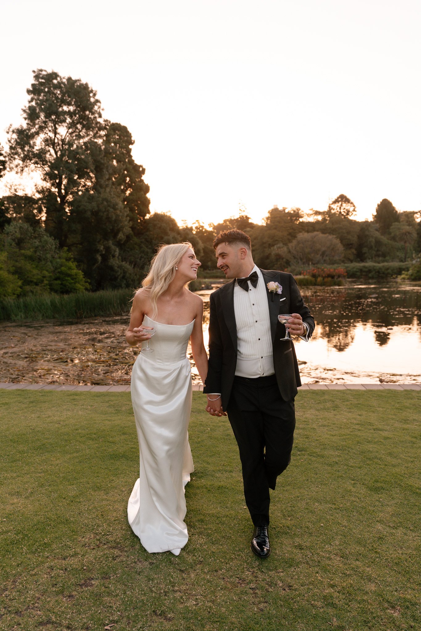 A bride and groom holding hands and smiling at each other outdoors during sunset, near a pond and trees.