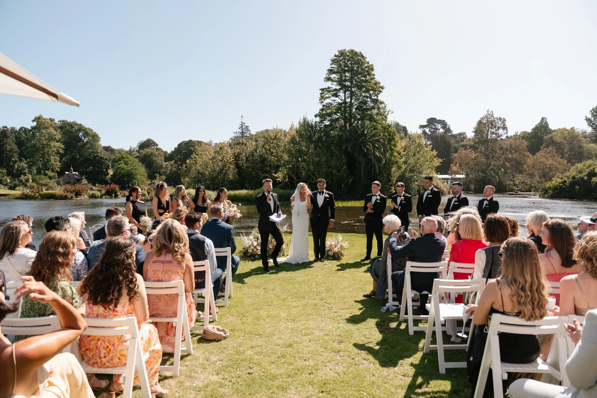 A wedding ceremony taking place outdoors on a grassy area near a body of water, with a crowd of guests seated on white chairs, a bride and groom standing together, and a officiant reading. The background features trees and a clear sky.