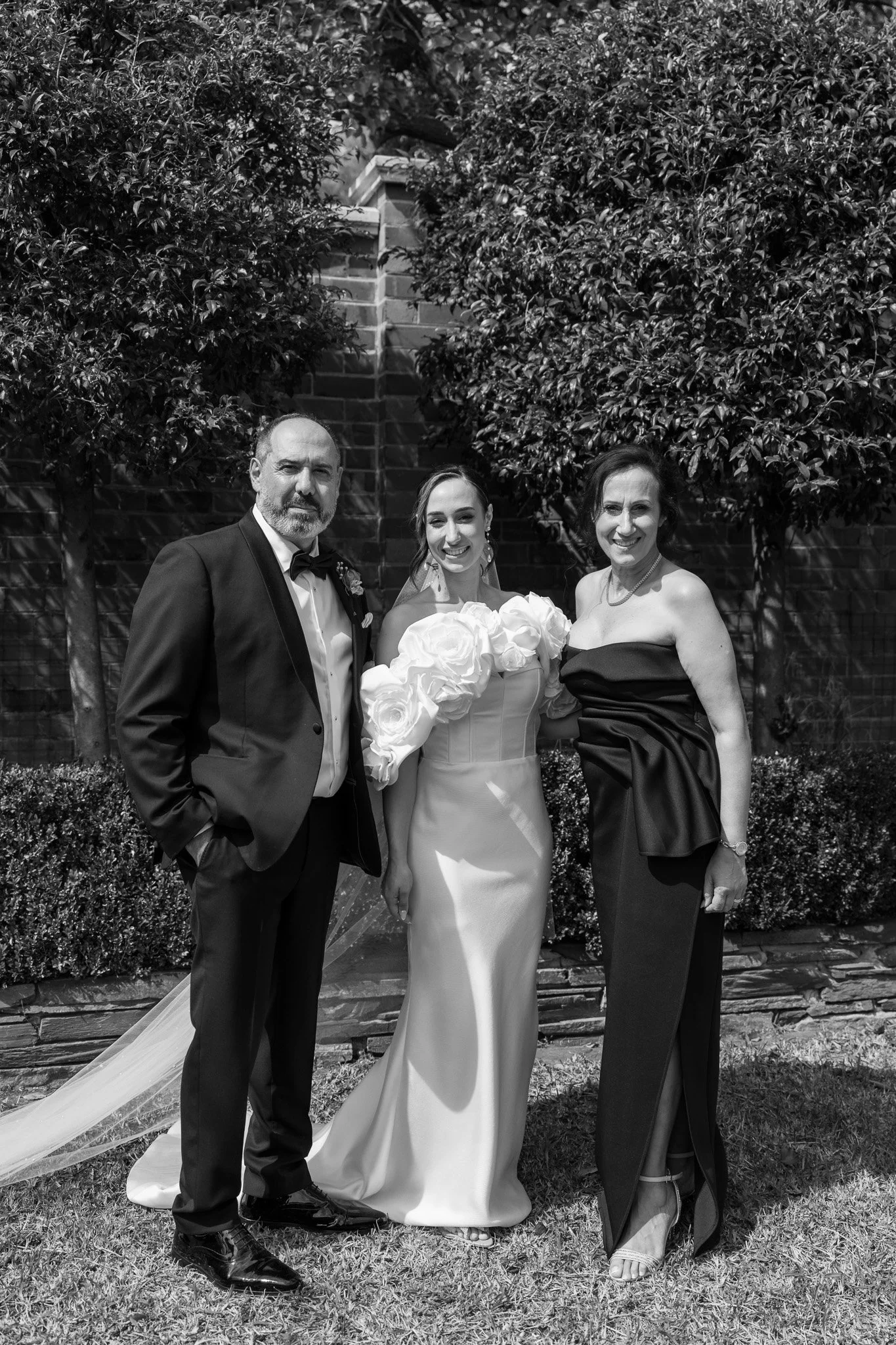 Black and white photo of three people at a wedding: a bride in a gown with large floral embellishments on her sleeves, a man in a tuxedo, and a woman in a strapless dress, standing outdoors in front of trees and a brick wall.