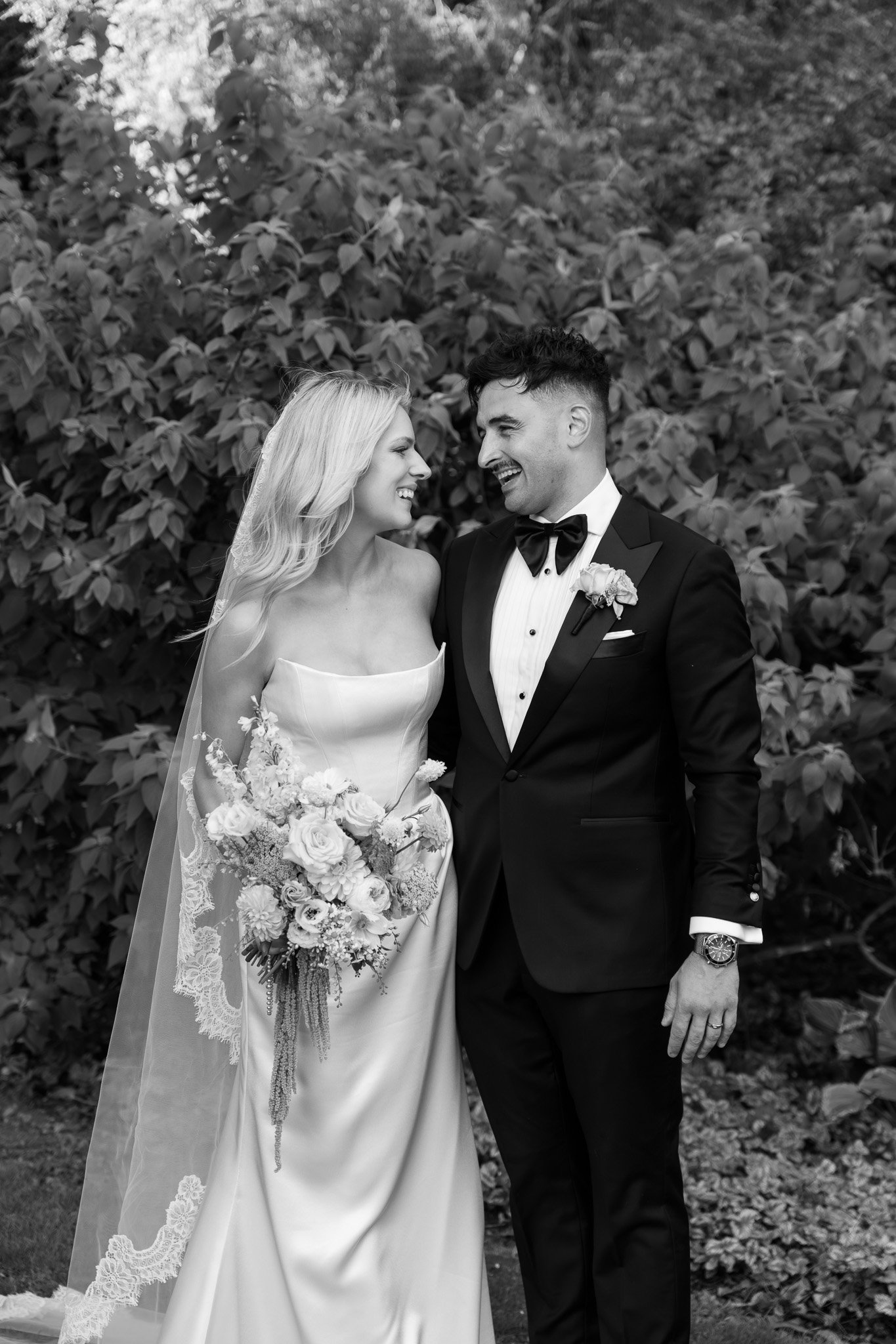 Black and white photo of a bride and groom standing close, smiling at each other outdoors with foliage in the background. The bride wears a strapless wedding dress and holds a bouquet of flowers, while the groom wears a tuxedo with a bow tie and a bo