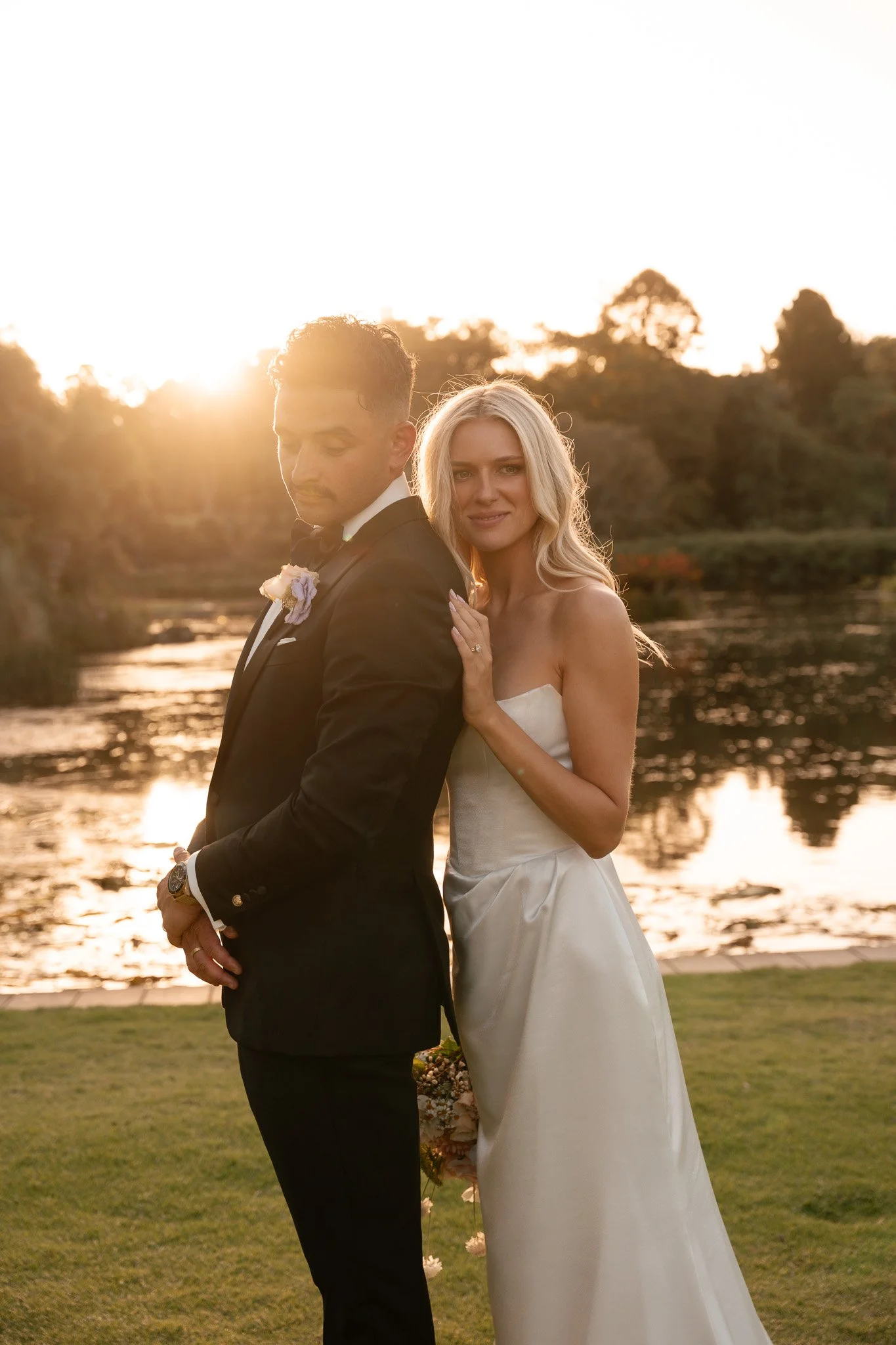 A bride and groom standing near a river during sunset, with the groom in a black tuxedo and the bride in a white strapless wedding dress, both looking peaceful.