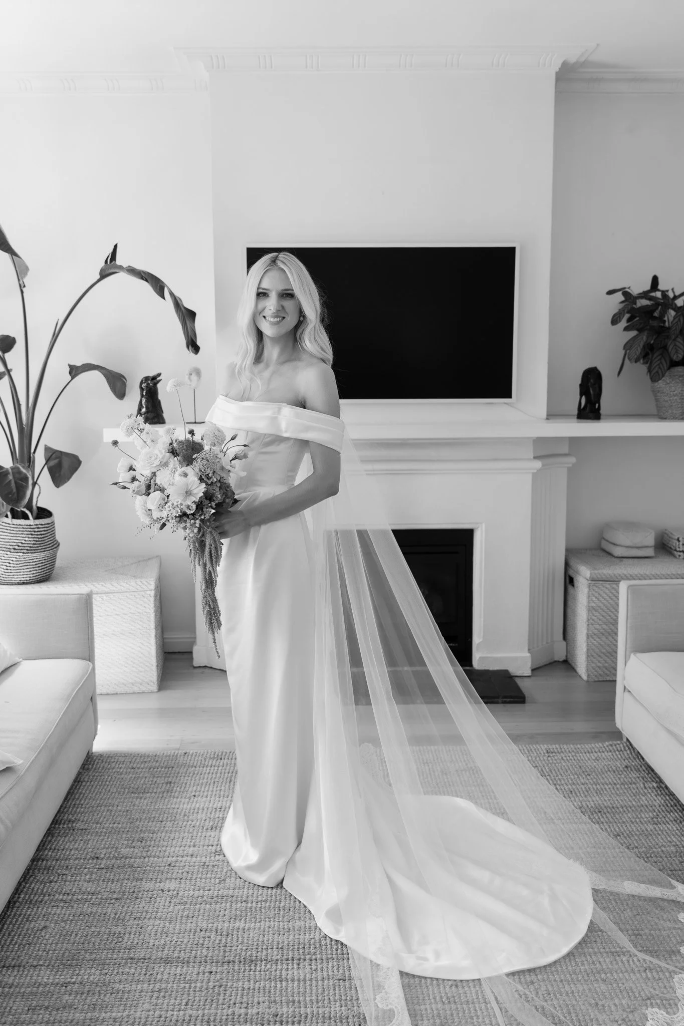 Black and white photo of a smiling bride in a wedding gown with a large bouquet, standing in a living room with a fireplace, TV, and decorative plants.