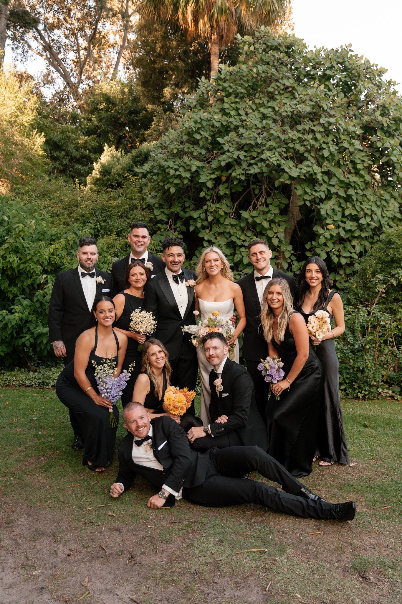 A group of people dressed formally, including a bride in a white gown, posing outdoors for a wedding photo with lush greenery and trees in the background.