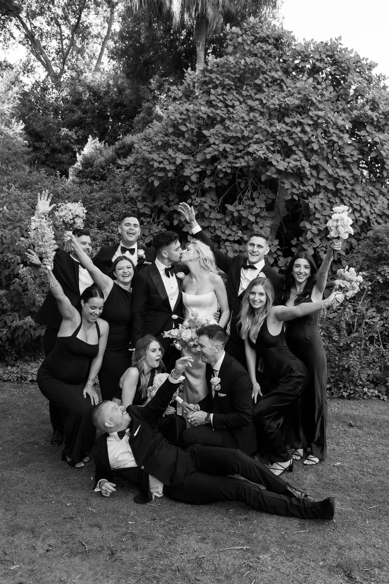 Black and white photo of a wedding party outdoors, with the bride and groom at the center surrounded by friends and family, some holding bouquets and celebrating.