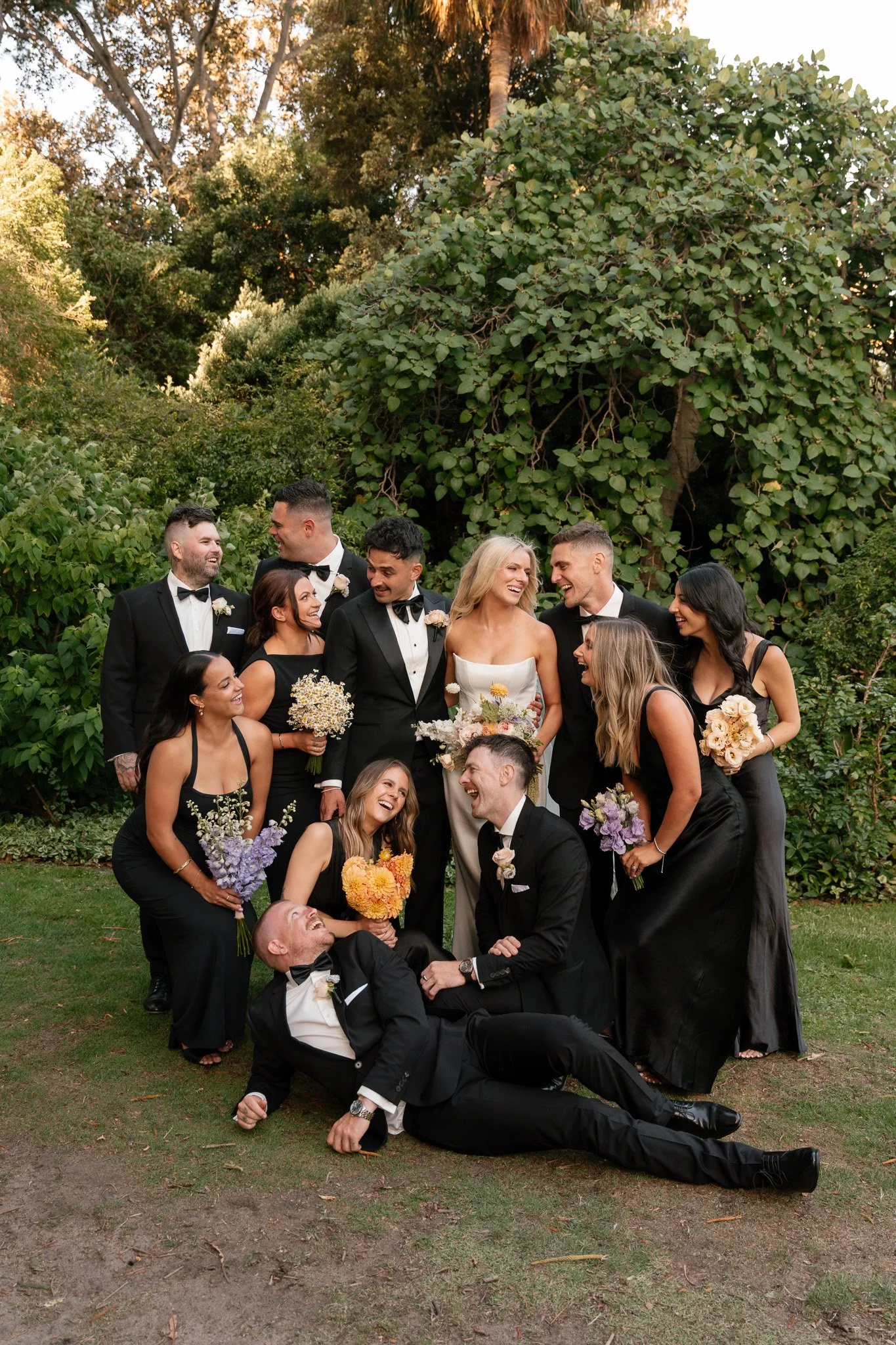 A group of people dressed in black formal wear, celebrating at a wedding outdoors, with trees and bushes in the background.